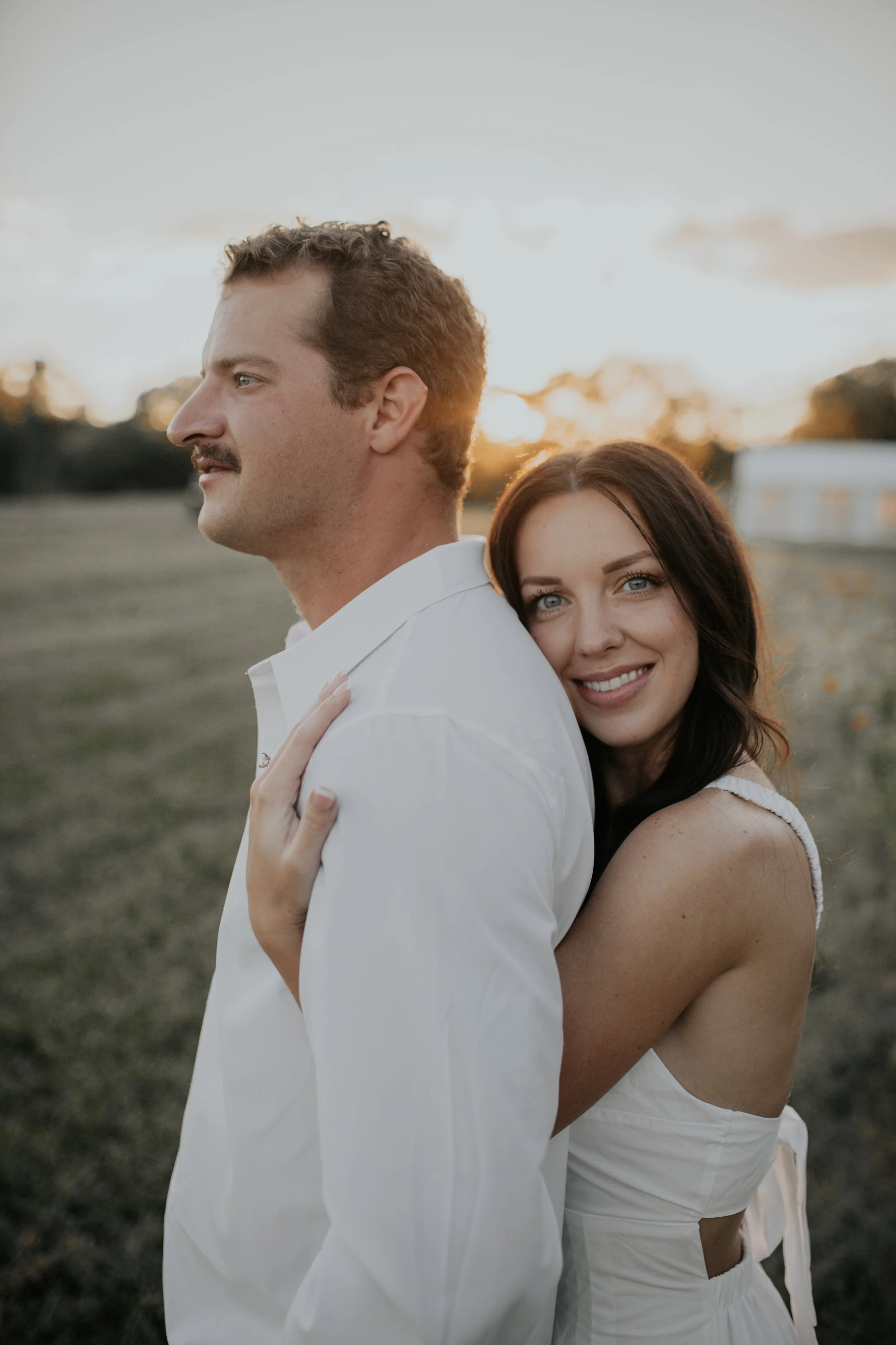 A couple in white clothes outdoors at sunset, with the woman hugging the man's back and smiling at the camera, the man looking to the left.