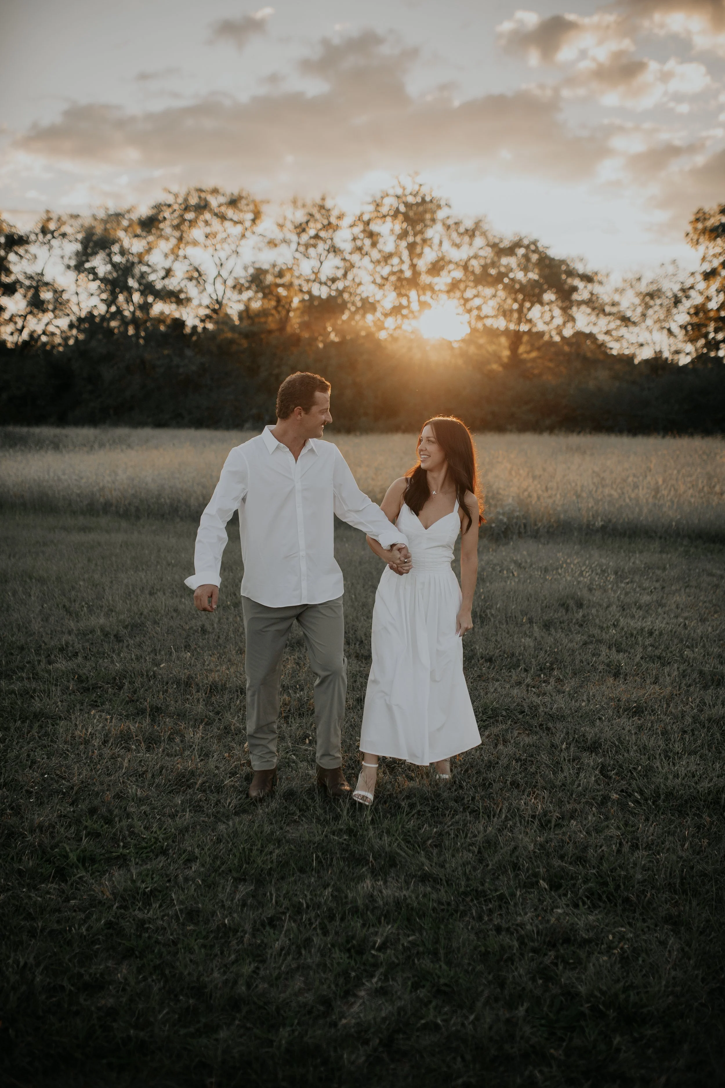 A couple walking hand in hand across a grassy field at sunset, with trees and a partly cloudy sky in the background.