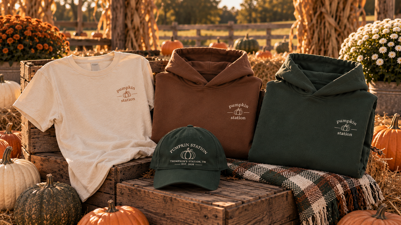 Set of pumpkins and fall apparel on a wooden display with a rustic background of trees and a fence, featuring a white t-shirt, two hoodies in brown and green, and a dark green cap, all branded with "Pumpkin Station."