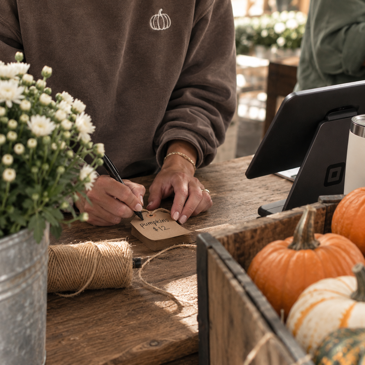 Person writing on a small brown paper bag that says "Pumpkins $12" at a farm stand. There is a bunch of white flowers, a spool of twine, and pumpkins on the rustic wooden counter.