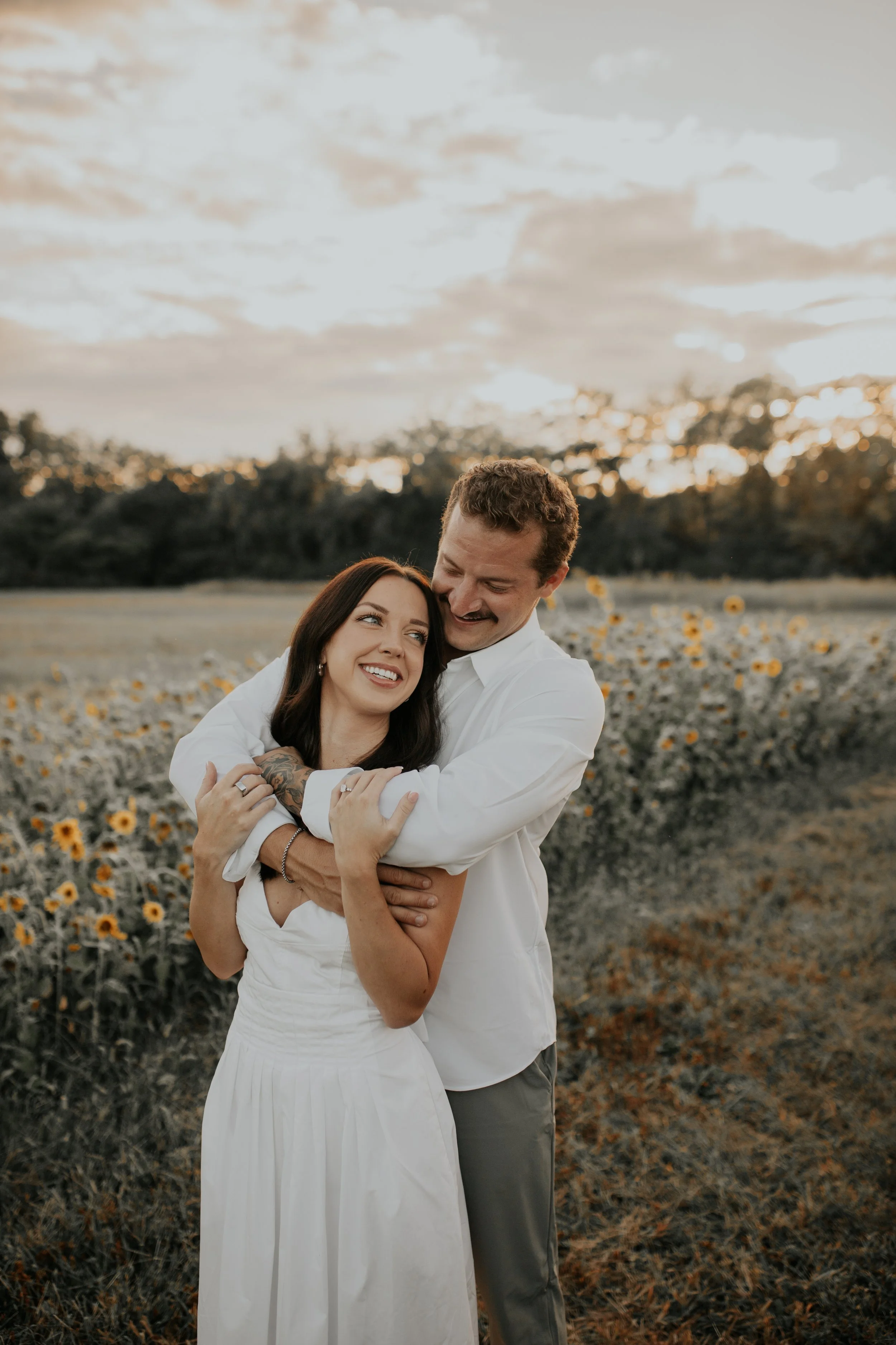 A happy couple embracing in a field of sunflowers at sunset, with the woman smiling at the man and the man looking down at her.