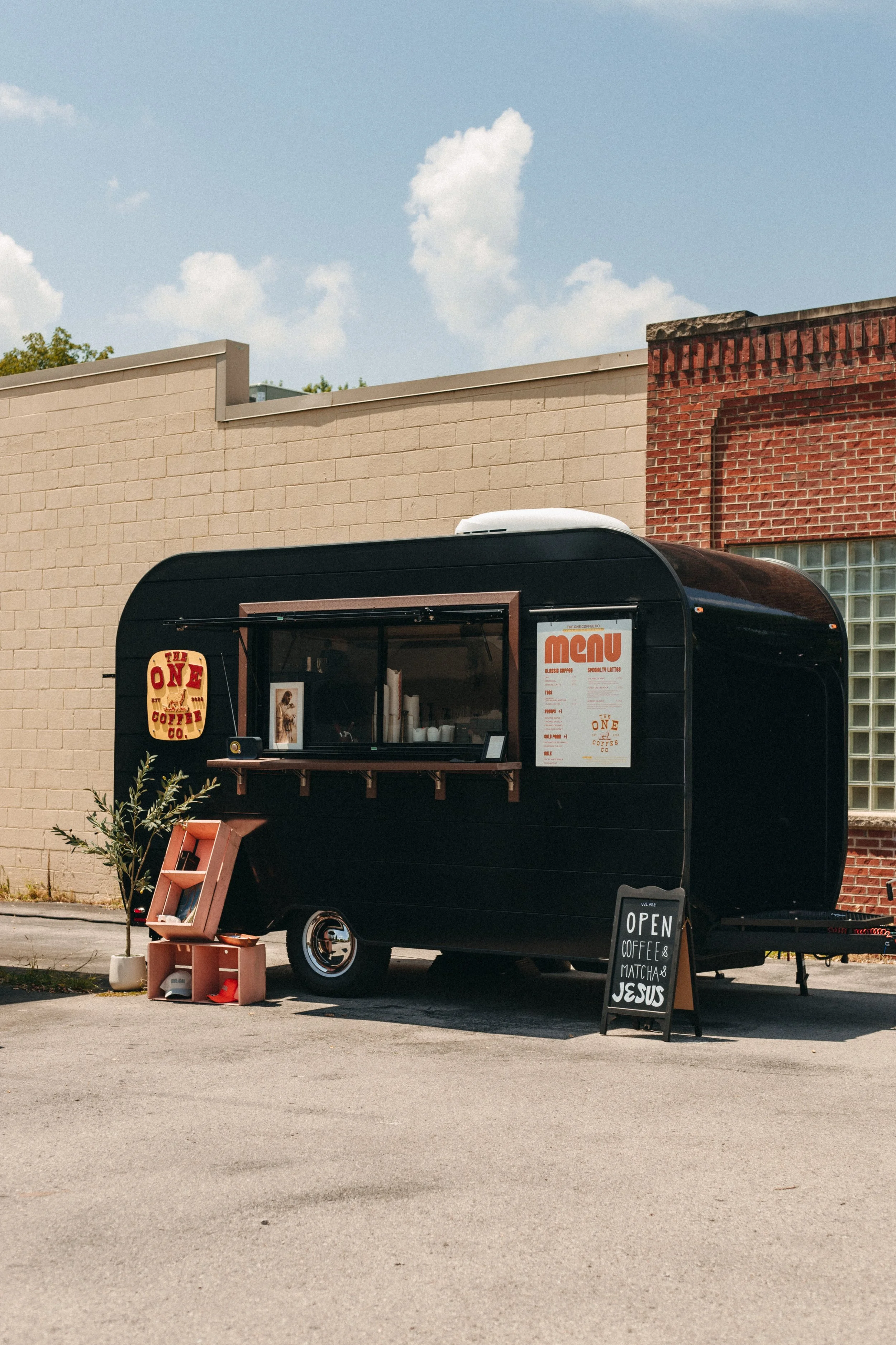 A black mobile coffee cart with a wooden menu sign, small shelves, and a chalkboard sign that reads 'Open Coffee & Matcha Jesus' is set up outside in a parking lot next to a beige brick building with a red brick section. There's a small table with a plant and some items in front of the cart, and a small sign on the ground. The background features a blue sky with scattered clouds.