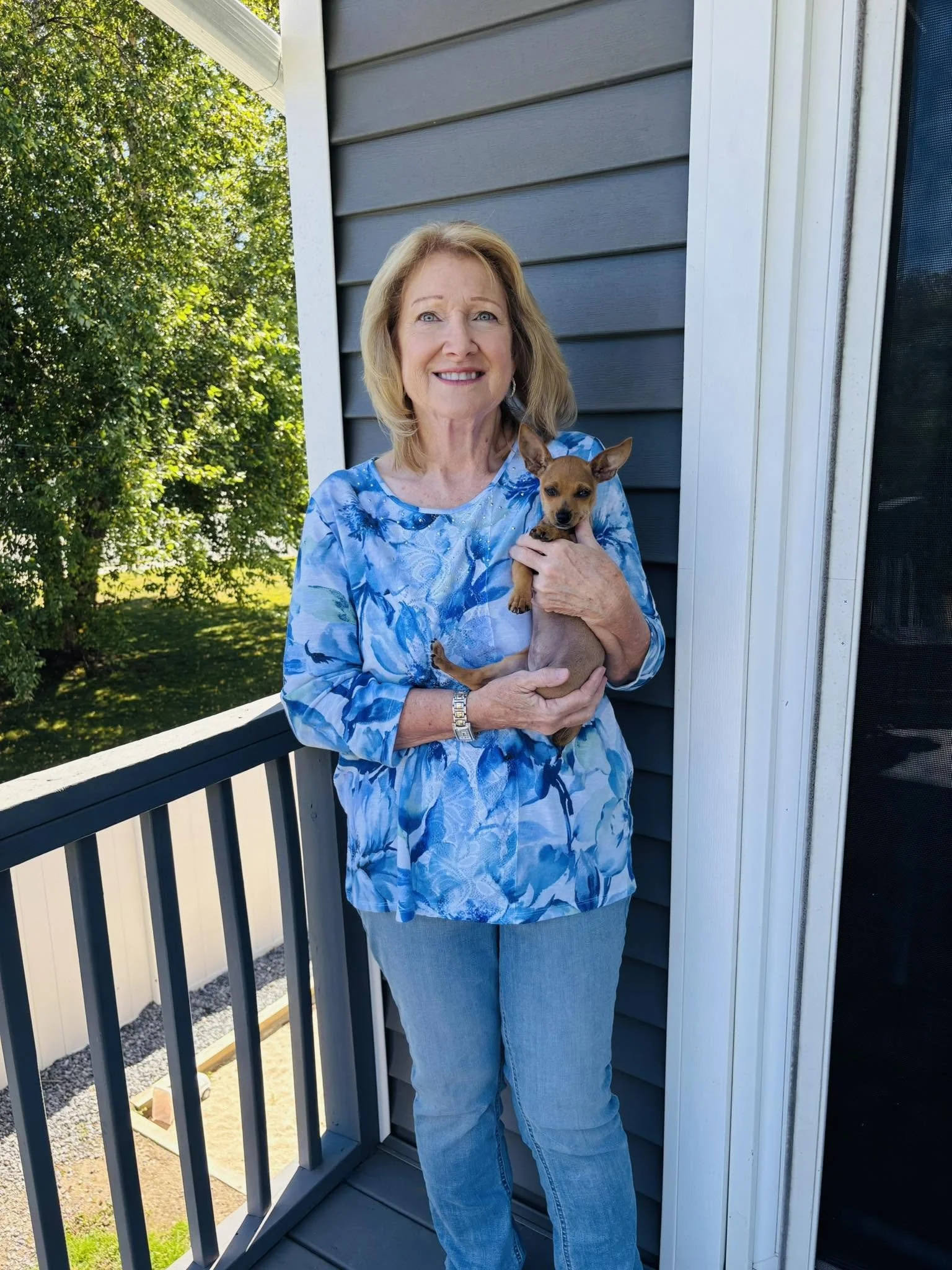 A woman with blonde hair in a blue patterned top and blue jeans holding a small brown dog on a balcony with a blue house exterior and trees in the background.
