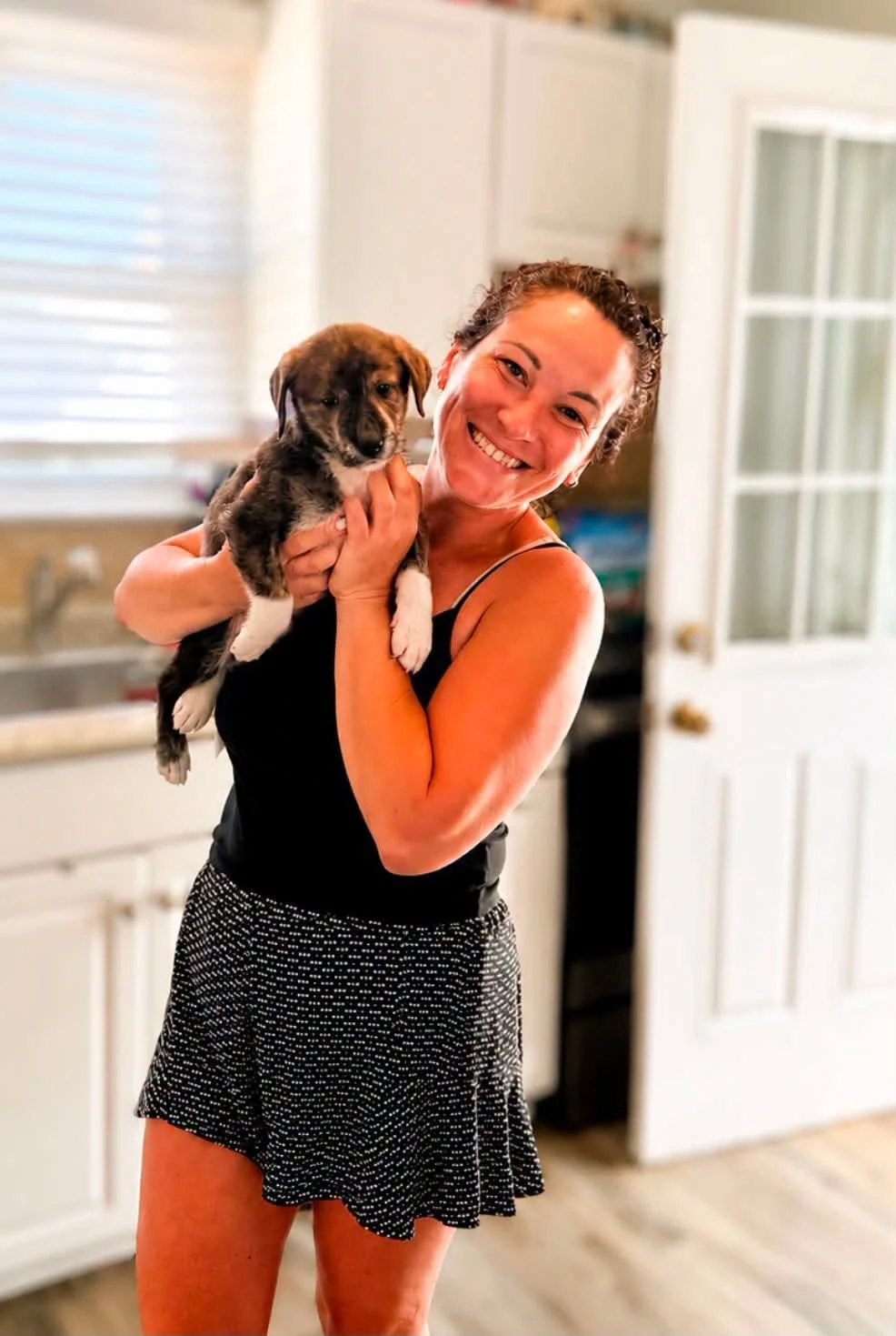 A woman smiling and holding a small puppy in a kitchen with white cabinets and a window.