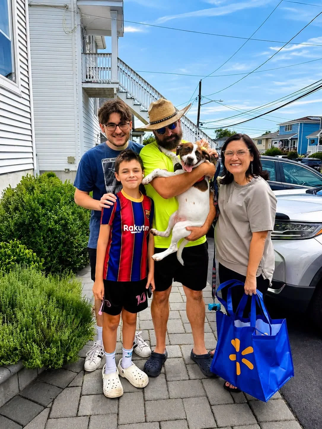 A happy family of four people and a dog standing on a sidewalk in a residential neighborhood with houses and cars in the background. The man in the center is holding a dog, and all are smiling at the camera.