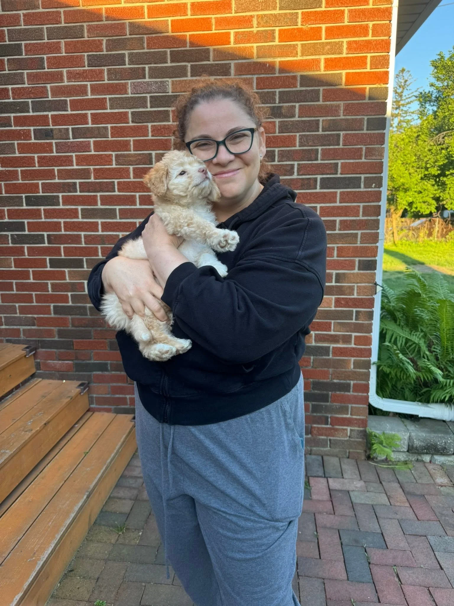 A woman with glasses and a black hoodie is holding a small, fluffy puppy with light-colored fur in front of a brick wall outside. The woman is smiling and standing on a paved walkway.