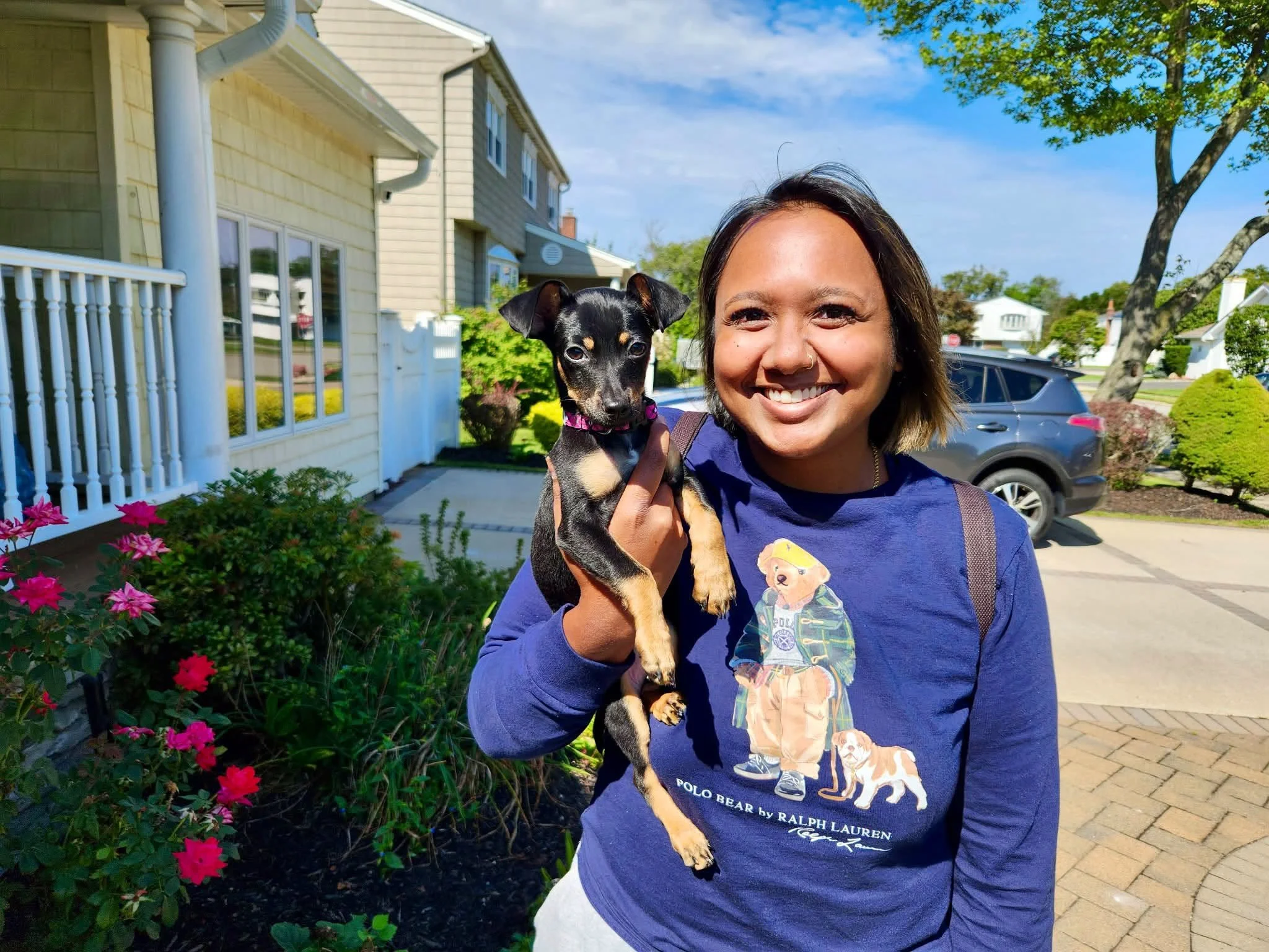 A woman smiling and holding a small black and tan puppy outdoors in front of a residential house with a white fence, pink flowers, green shrubs, and a large tree, under a partly cloudy blue sky.