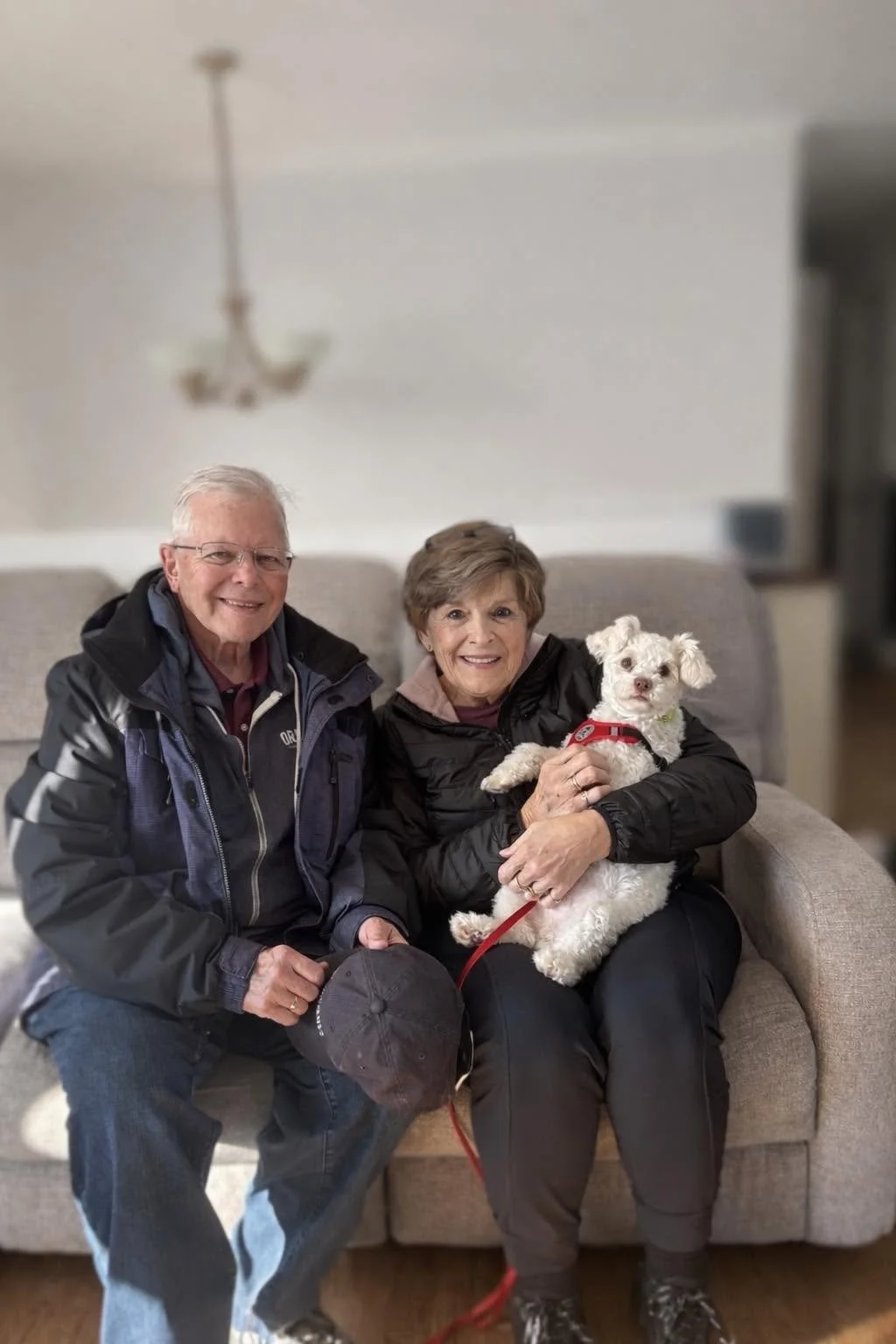 An elderly man and woman sitting on a beige couch indoors, smiling at the camera, with the woman holding a small white dog with floppy ears and a red harness.