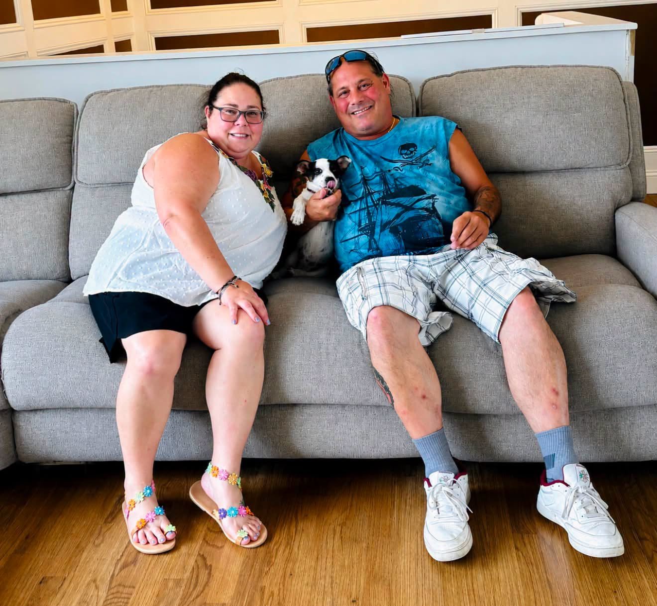 A woman and a man sitting on a gray couch, smiling, with a small black and white puppy between them. The woman is wearing glasses, a white sleeveless top, and black shorts. The man is wearing a blue T-shirt with a skull and pirate ship graphic, plaid shorts, and sneakers. The living room has wood flooring and patterned wall panels in the background.