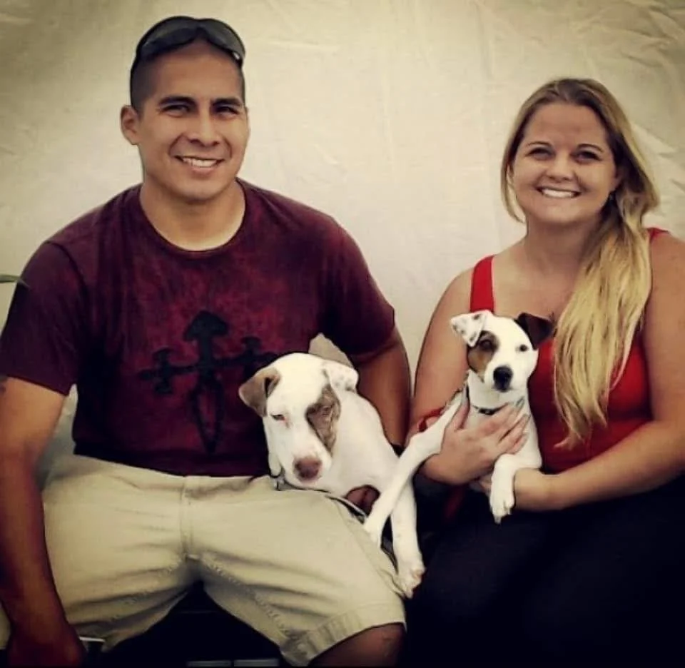 A smiling man and woman sitting side by side, each holding a dog, in front of a neutral background.