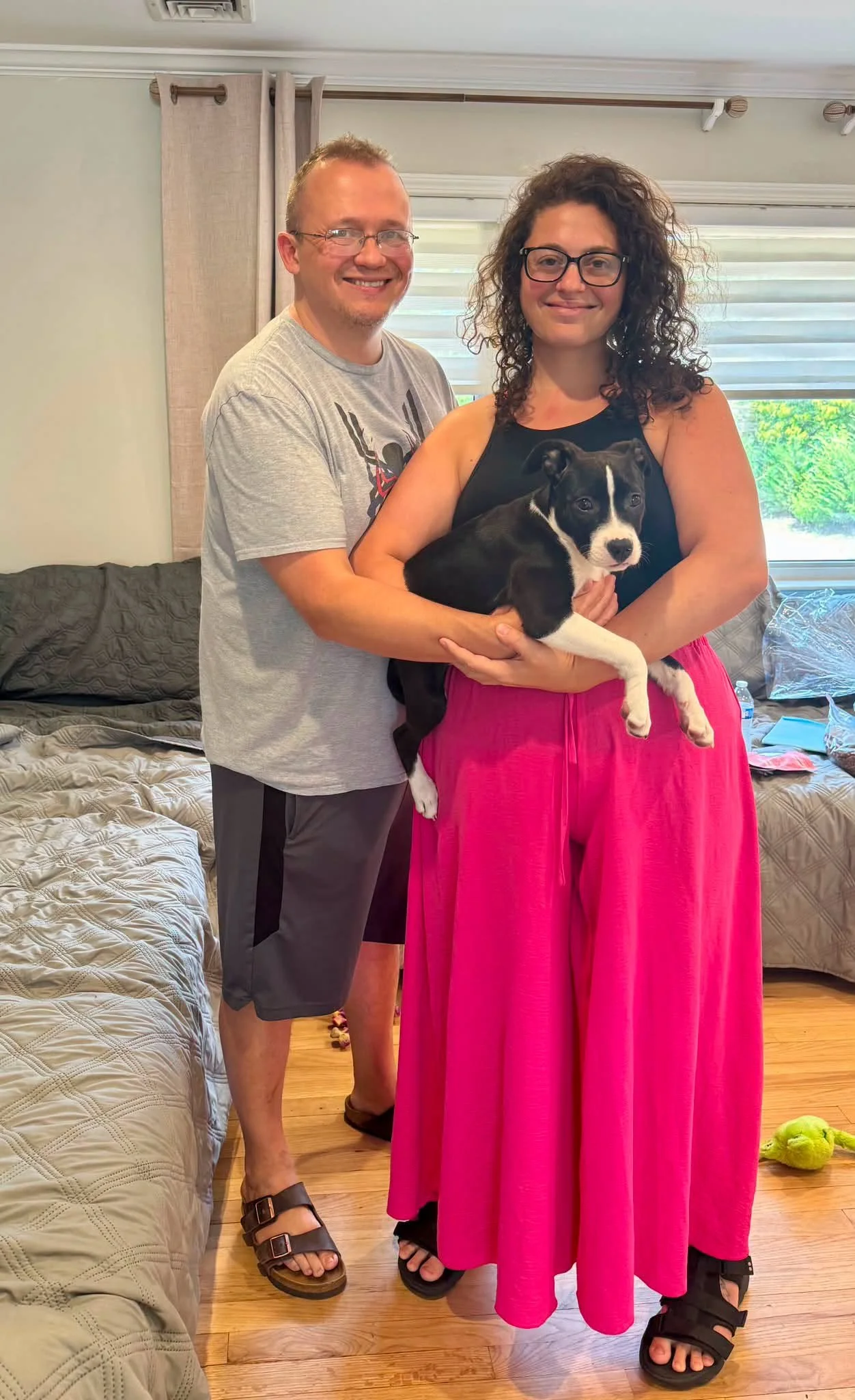 Two people, a man and a woman, standing indoors with a small black and white dog. The man is smiling and wearing a gray t-shirt, shorts, and sandals. The woman is holding the dog, wearing glasses, a black top, a bright pink skirt, and black sandals. The room has wooden floors, a bed, and a window with blinds and curtains.