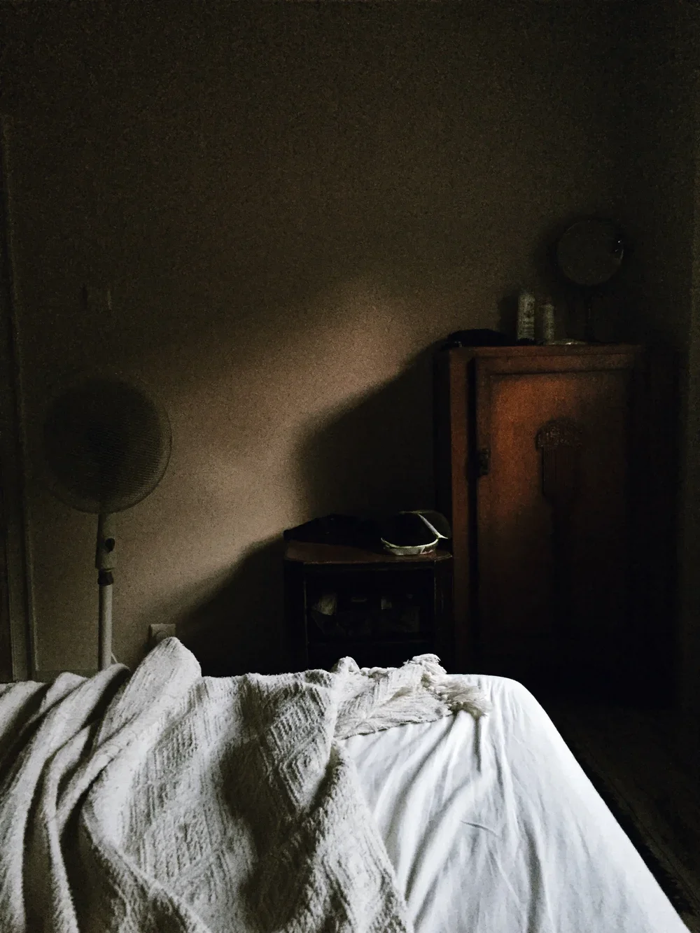 A dimly lit bedroom with a bed, a white textured blanket, an alarm clock, a wooden cabinet, a standing fan, and some bottles on the cabinet.