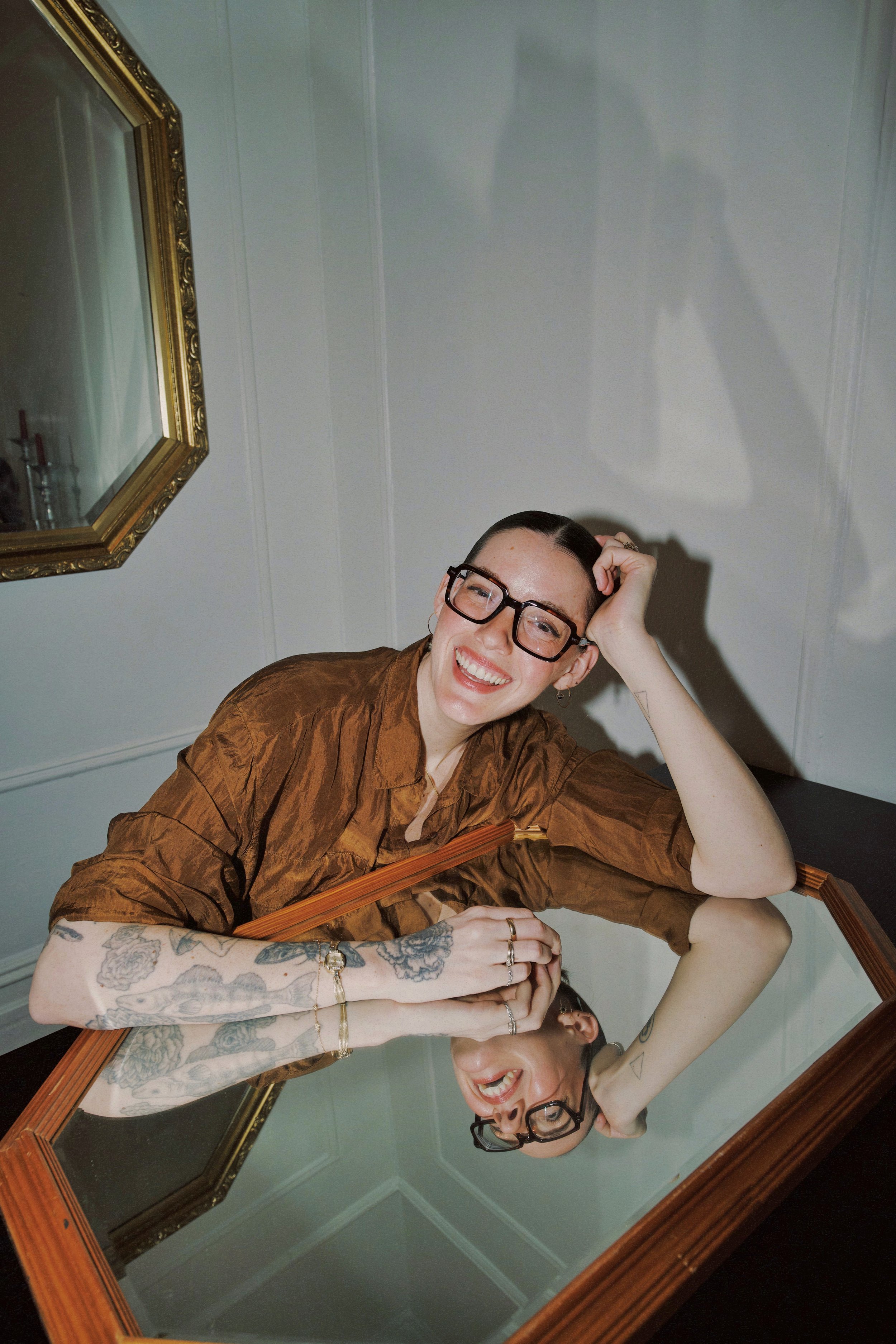 A smiling person with glasses and tattoos, resting their head on one hand, sits at a reflective surface with a framed mirror on the wall. The photo is mirrored, showing the person and surroundings upside down.