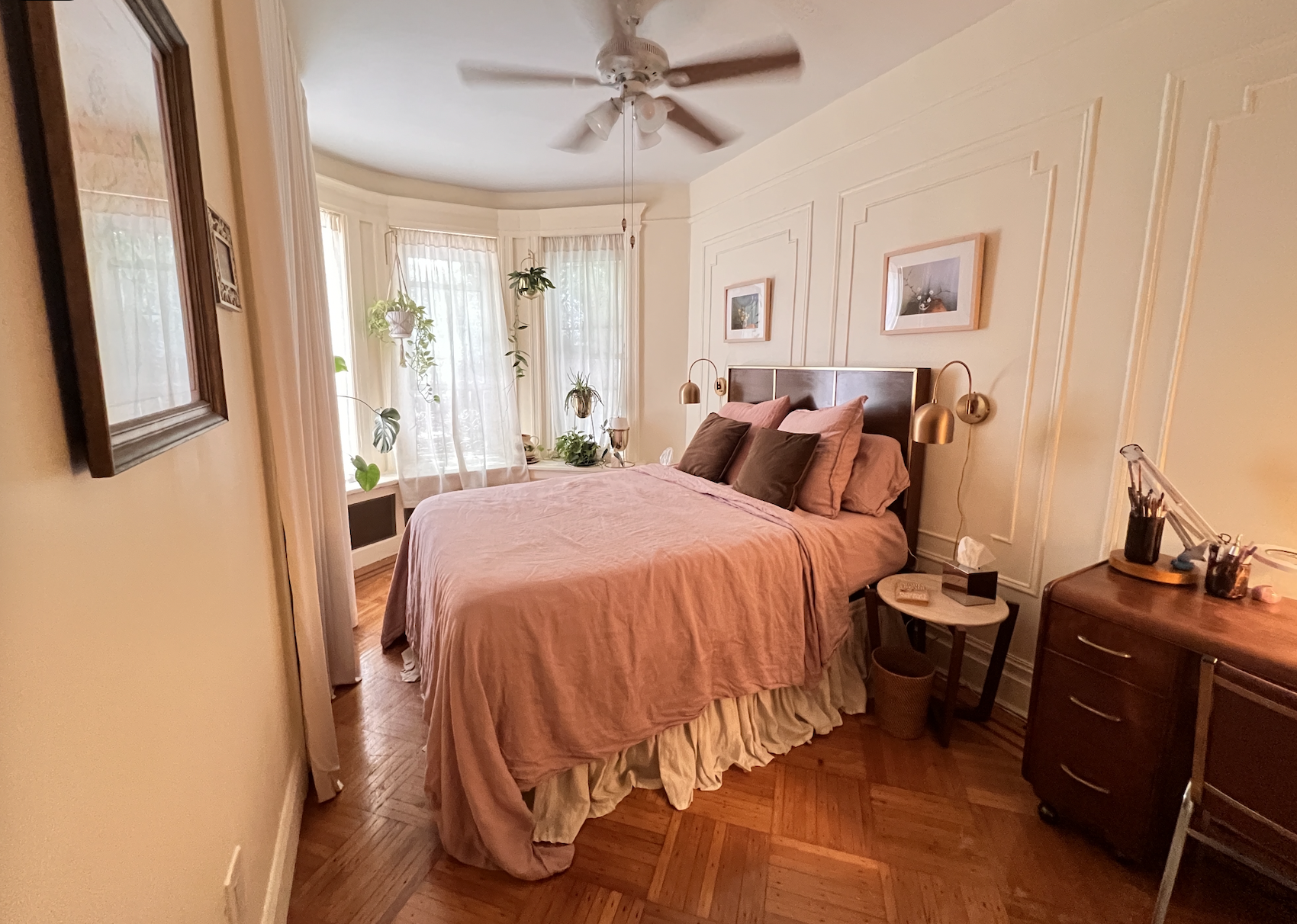 A cozy bedroom with a double bed covered in pink bedding and brown pillows. There are two hanging gold lamps on either side of the bed, a nightstand with a tissue box and small items, and a wooden dresser with art supplies. The room features a bay window with sheer curtains, several potted plants, and framed artwork on the walls. A ceiling fan is above, and hardwood floors complete the warm, inviting atmosphere.
