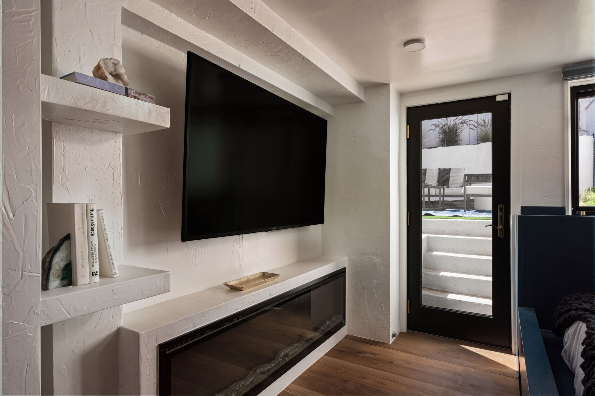 A living room corner featuring a wall-mounted flat-screen television, white textured wall with built-in shelves holding books and decorative items, a small fireplace below, and a glass door leading outside to a patio with furniture and outdoor plants