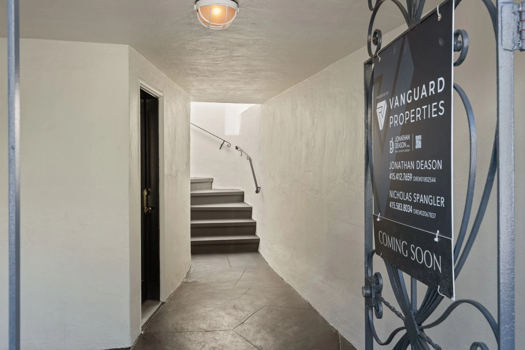 View of a stairwell with beige walls and stairs leading upward, partially visible, in a residential building. A black metal sign on a stand is in the foreground listing real estate agents and upcoming property availability.