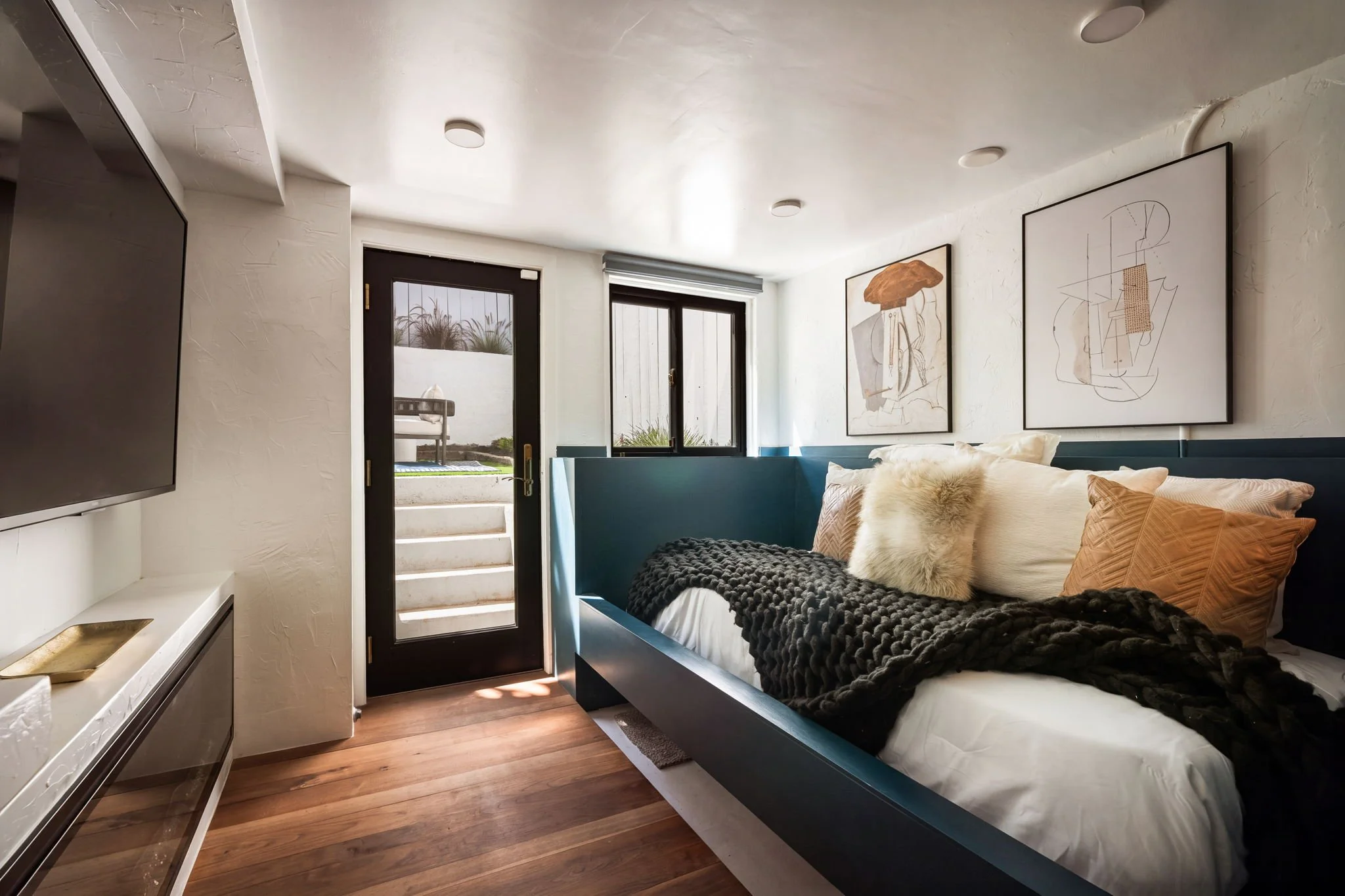 A cozy bedroom featuring a dark frame bed with white bedding, a furry pillow, orange pillow, and a textured blanket. The room has a large window and a glass door leading outside, with framed abstract art on the wall. The floor is wooden, and there ar