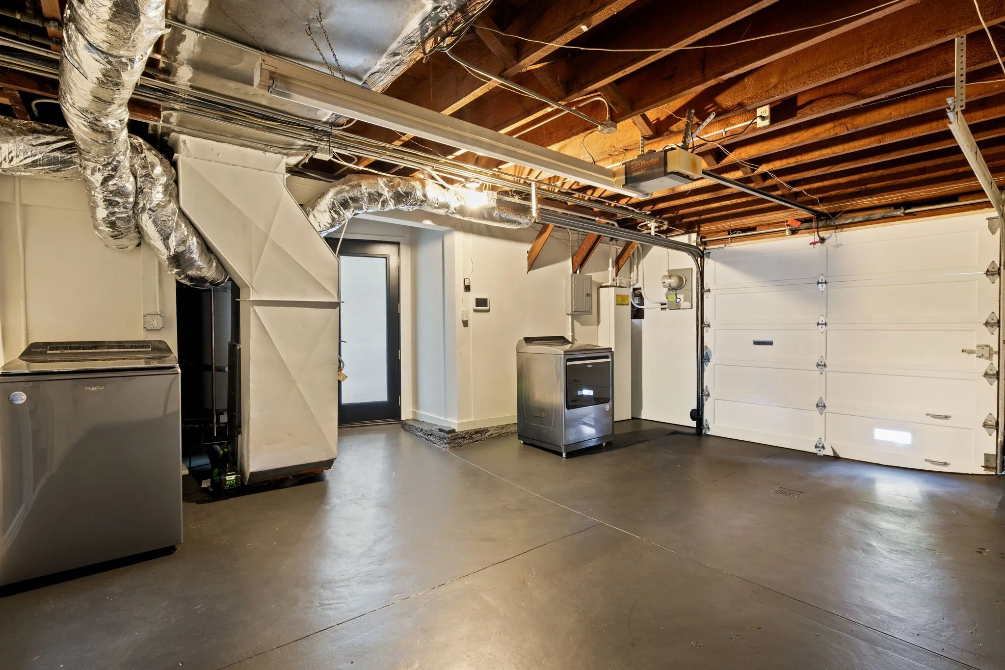 An unfinished garage with a cement floor, exposed wooden ceiling beams, and various HVAC and electrical components along the ceiling. There is a white garage door on the right and a black door with a window on the left, with laundry appliances and a 