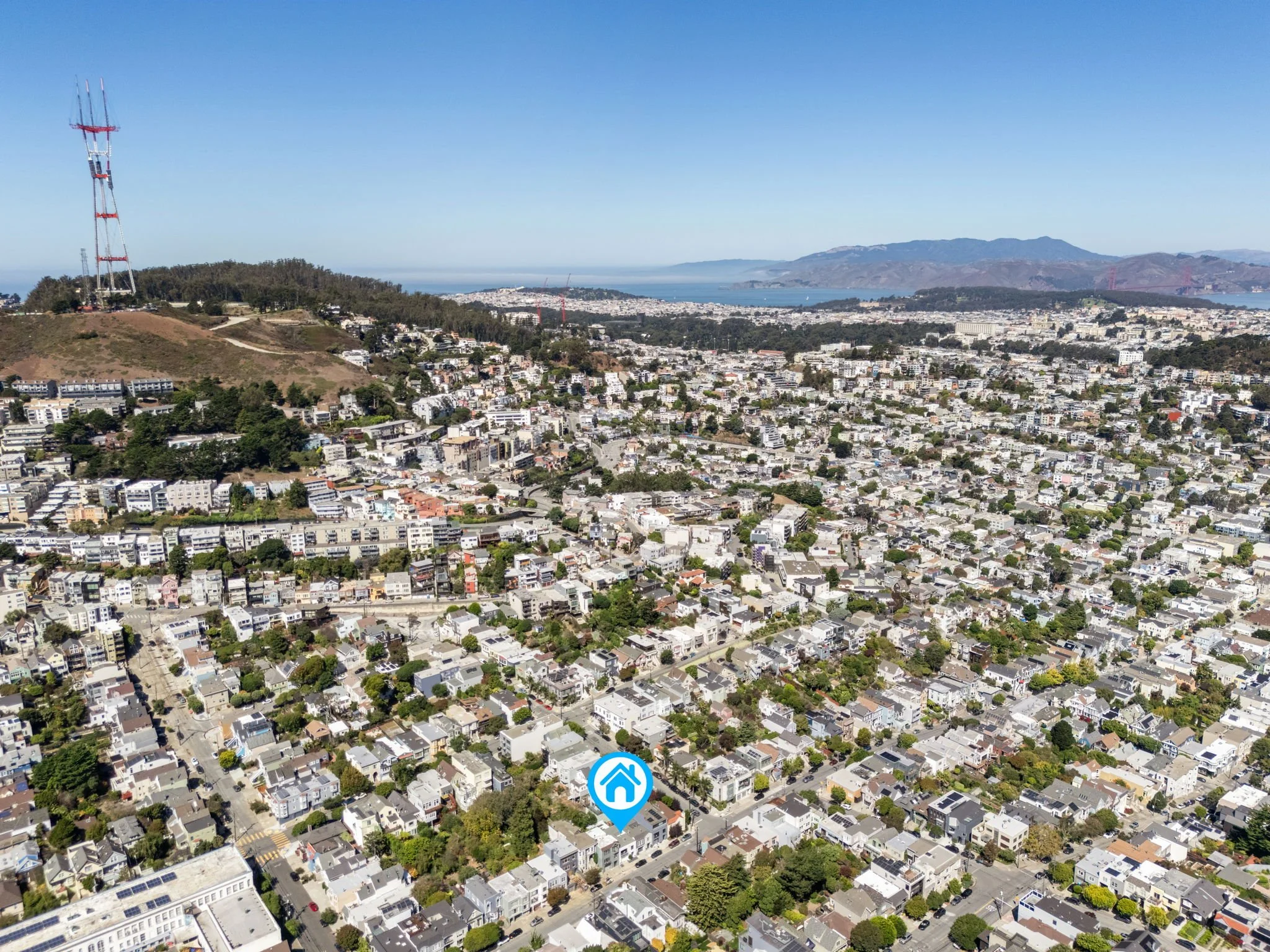 Aerial view of a densely populated city with a mix of residential and commercial buildings, hills with radio towers, and mountains in the background under a clear blue sky.