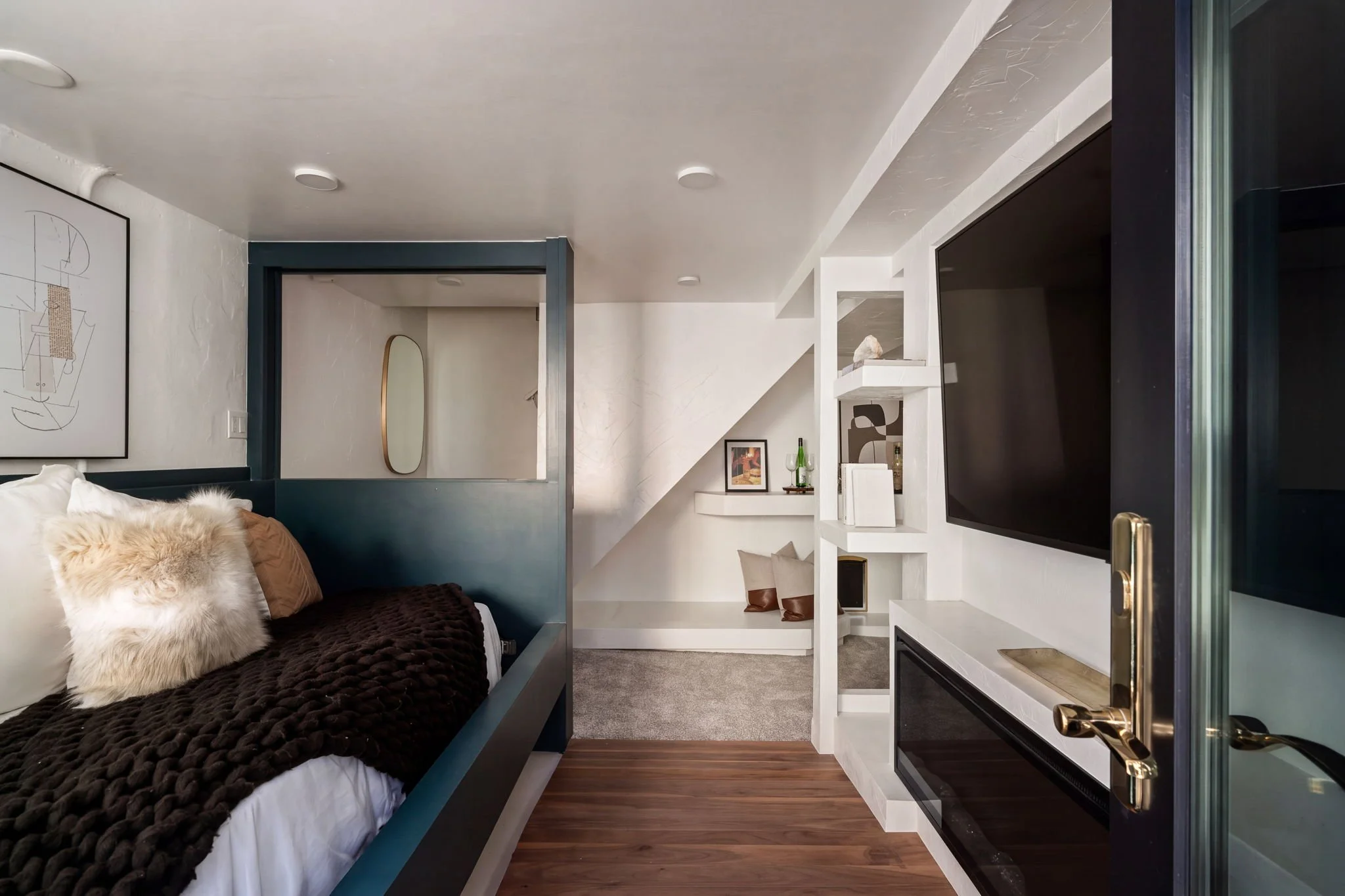 Interior of a modern living space with a bed on the left, decorative pillows, a geometric artwork on the wall, and a flat-screen TV on the right wall. There are also shelves with decorative items and a small countertop with a wine bottle and glasswar