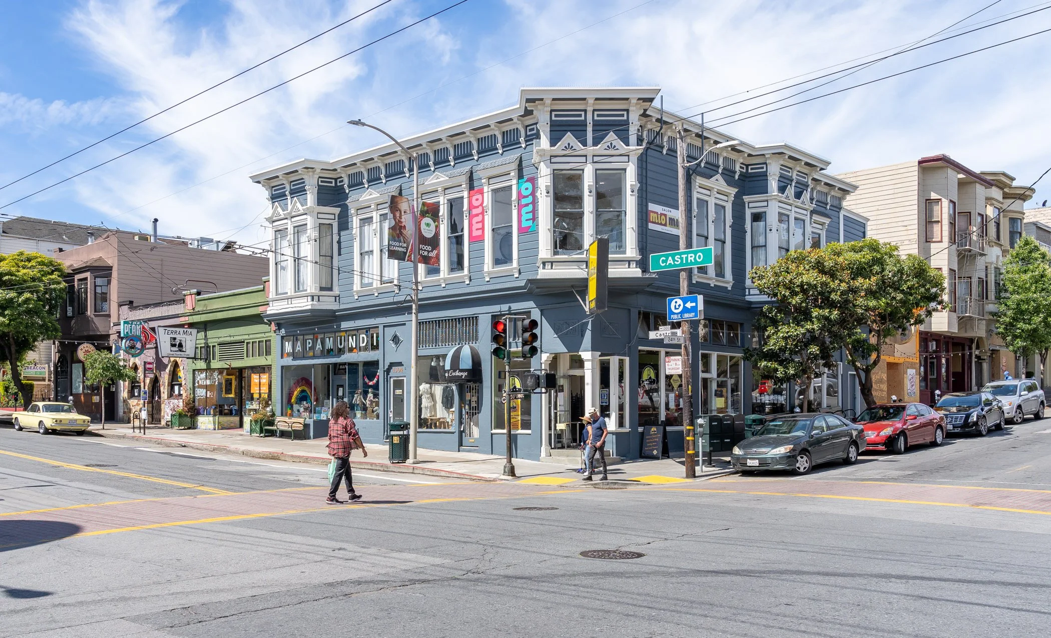 Street scene featuring a blue Victorian-style building with shops on the ground floor, including a bookstore called 'Mapamundi'. There are pedestrians walking across the street, cars parked along the curb, and a street sign indicating 'Castro'. Overh