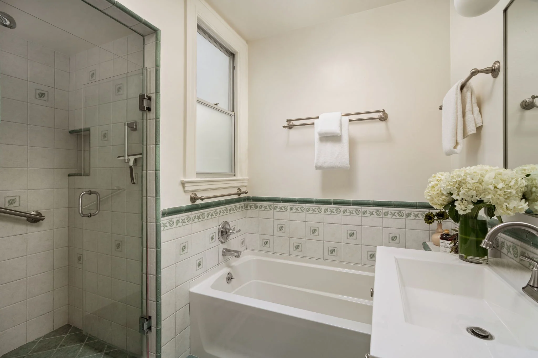 A bathroom with a shower enclosure on the left, tiled walls with a decorative border, a window above a bathtub on the right, and a sink with a faucet in the foreground, along with a towel rack with white towels and a flower arrangement on the sink.