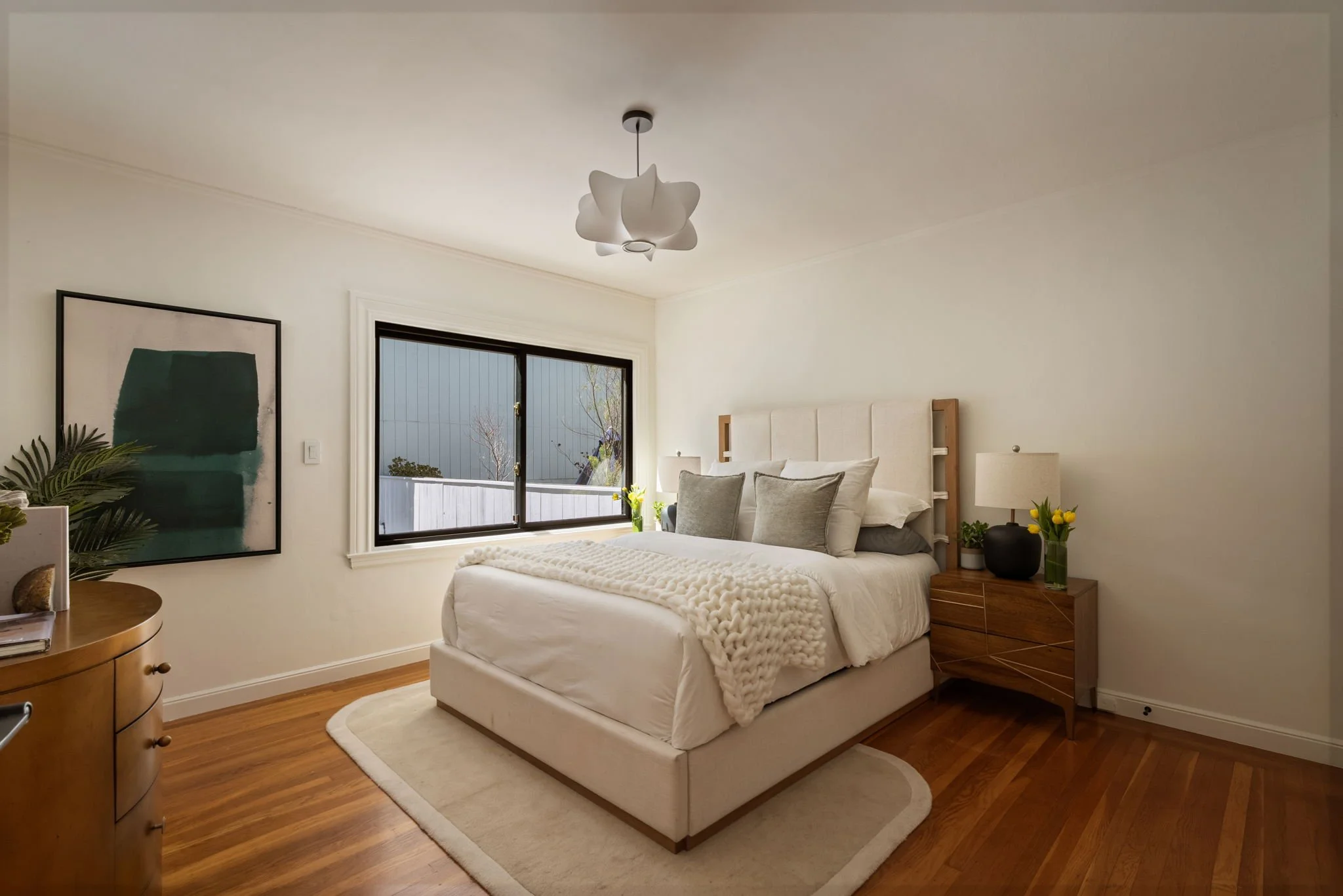 A cozy bedroom with a white bed, gray pillows, a wooden nightstand with a black vase and yellow flowers, a window with black frame, modern ceiling light, and artwork on the wall.
