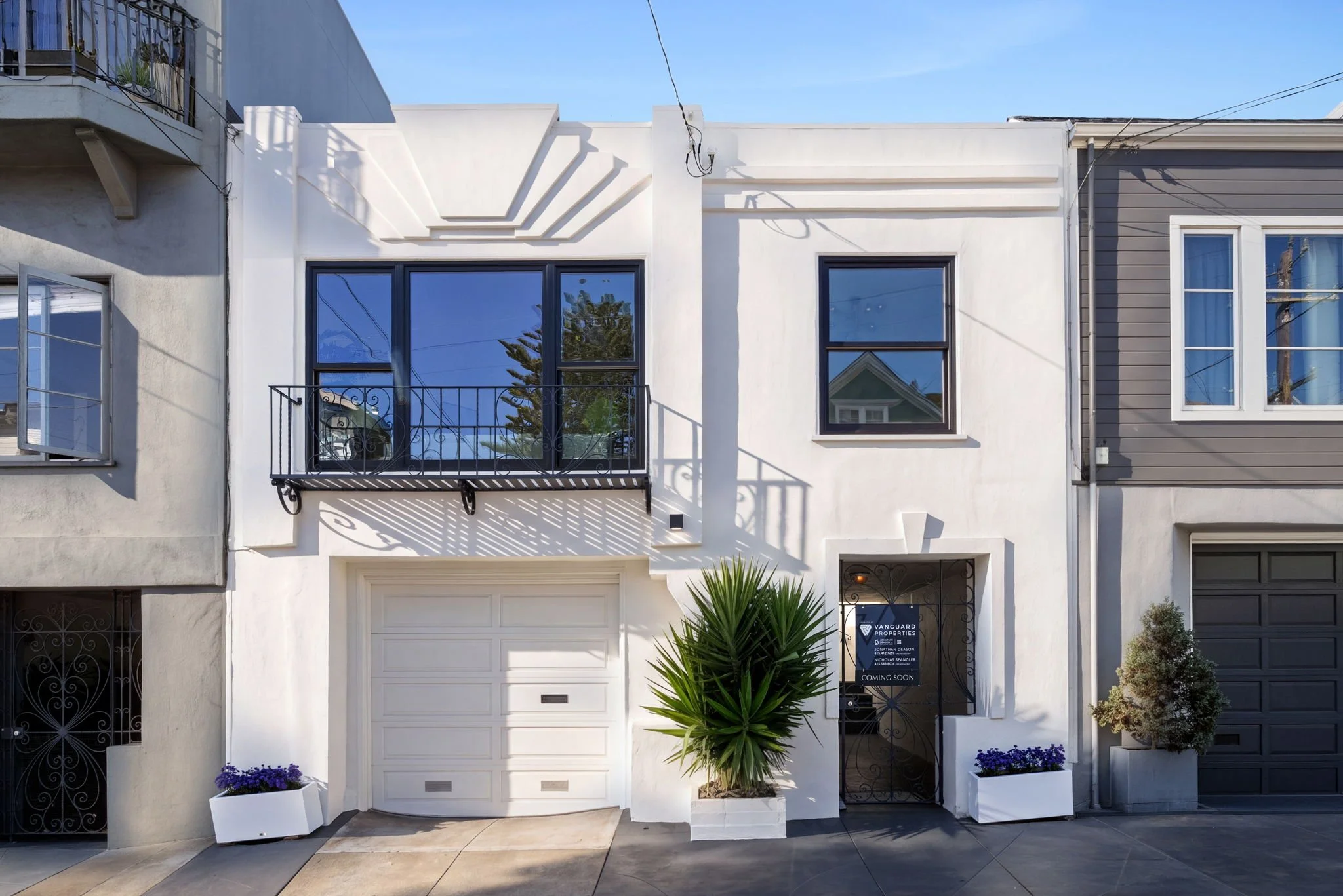 Modern white townhouse with large windows, a small balcony, black railings, and a nearby black fence. Two white planters with purple flowers and a green palm plant in front. Sign on the door indicates property is coming soon.