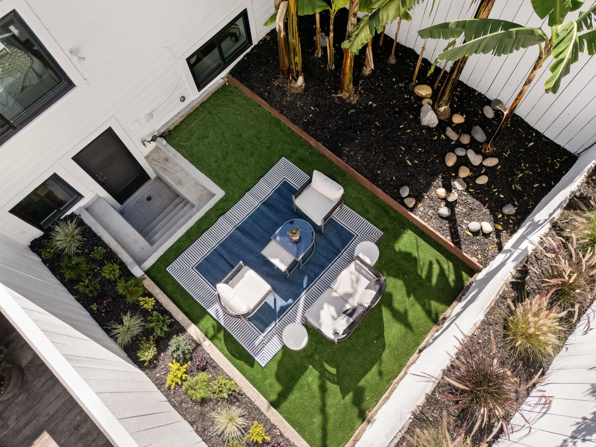 An aerial view of a backyard patio with a blue and white striped rug, four white armchairs, and round white side tables. There is a small patch of artificial grass and a fenced garden bed with banana plants and rocks.