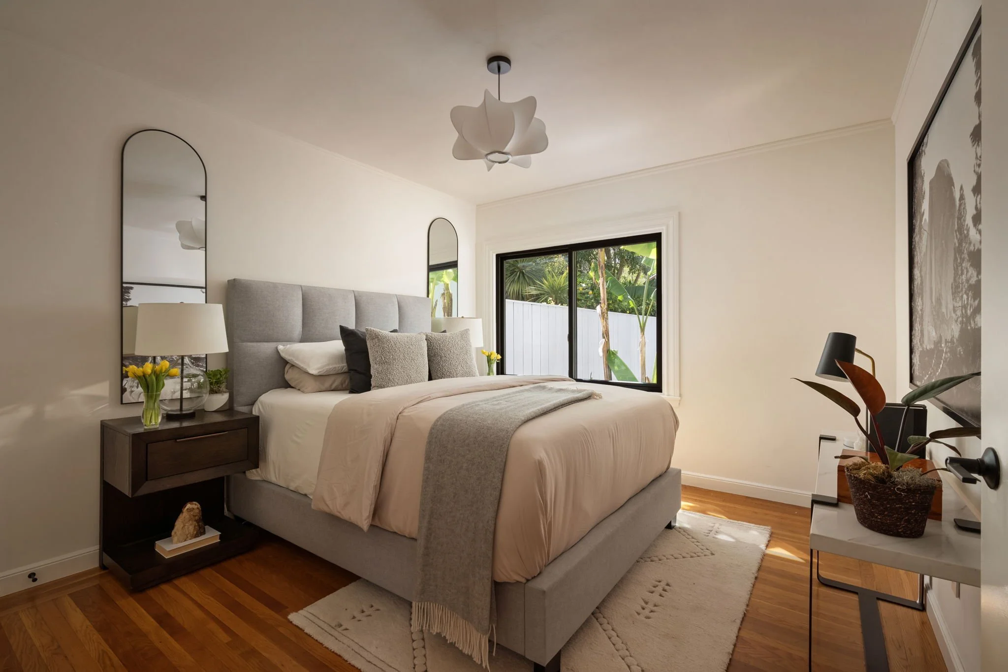 A bedroom with a bed, nightstand, lamps, large window showing greenery outside, and modern decor.