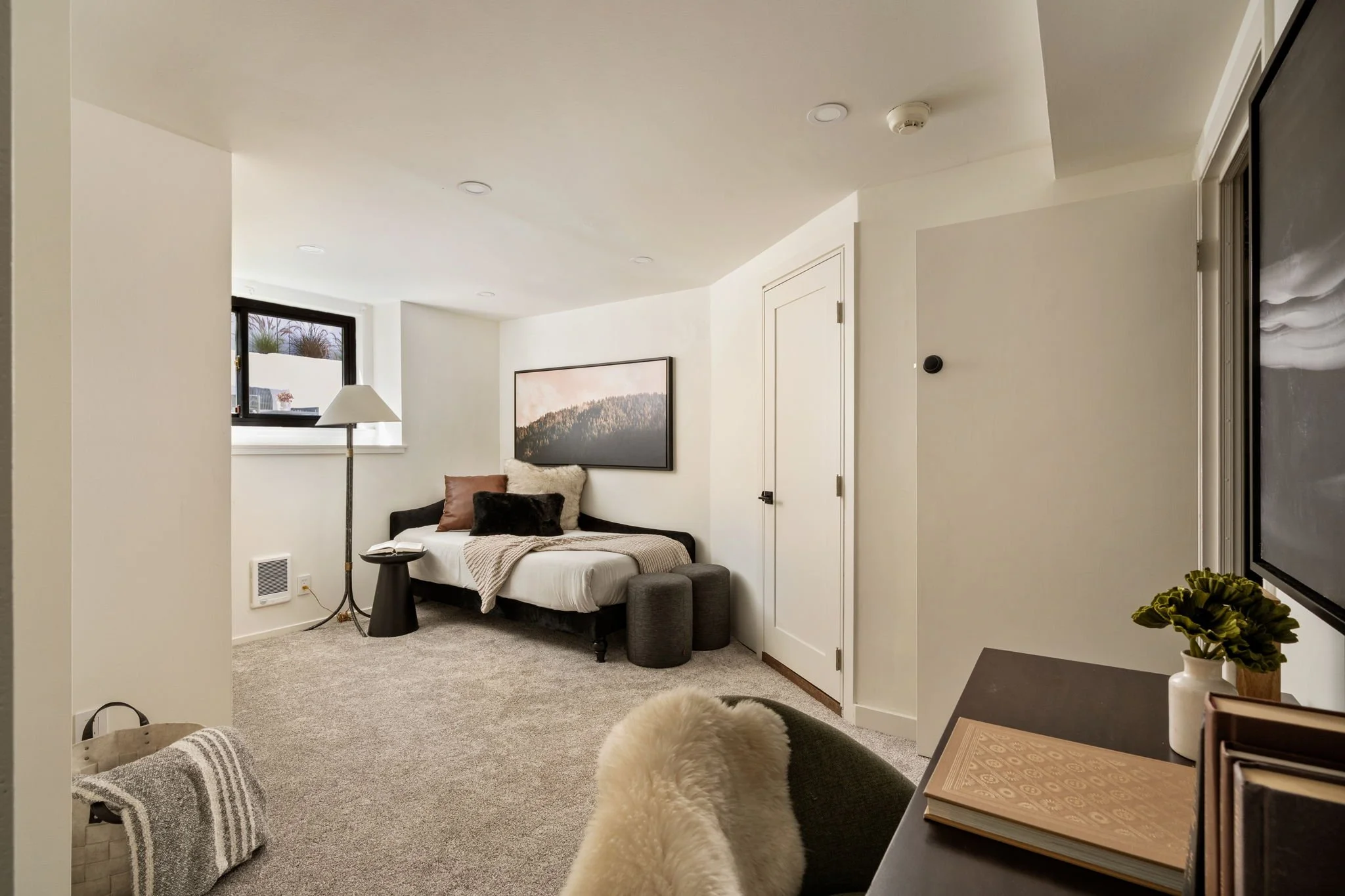 A cozy bedroom with a bed covered with a beige blanket and several pillows, a black side table, a standing lamp, a framed landscape picture on the wall, a window with a view of a balcony, and some indoor plants. The room has a neutral color palette w