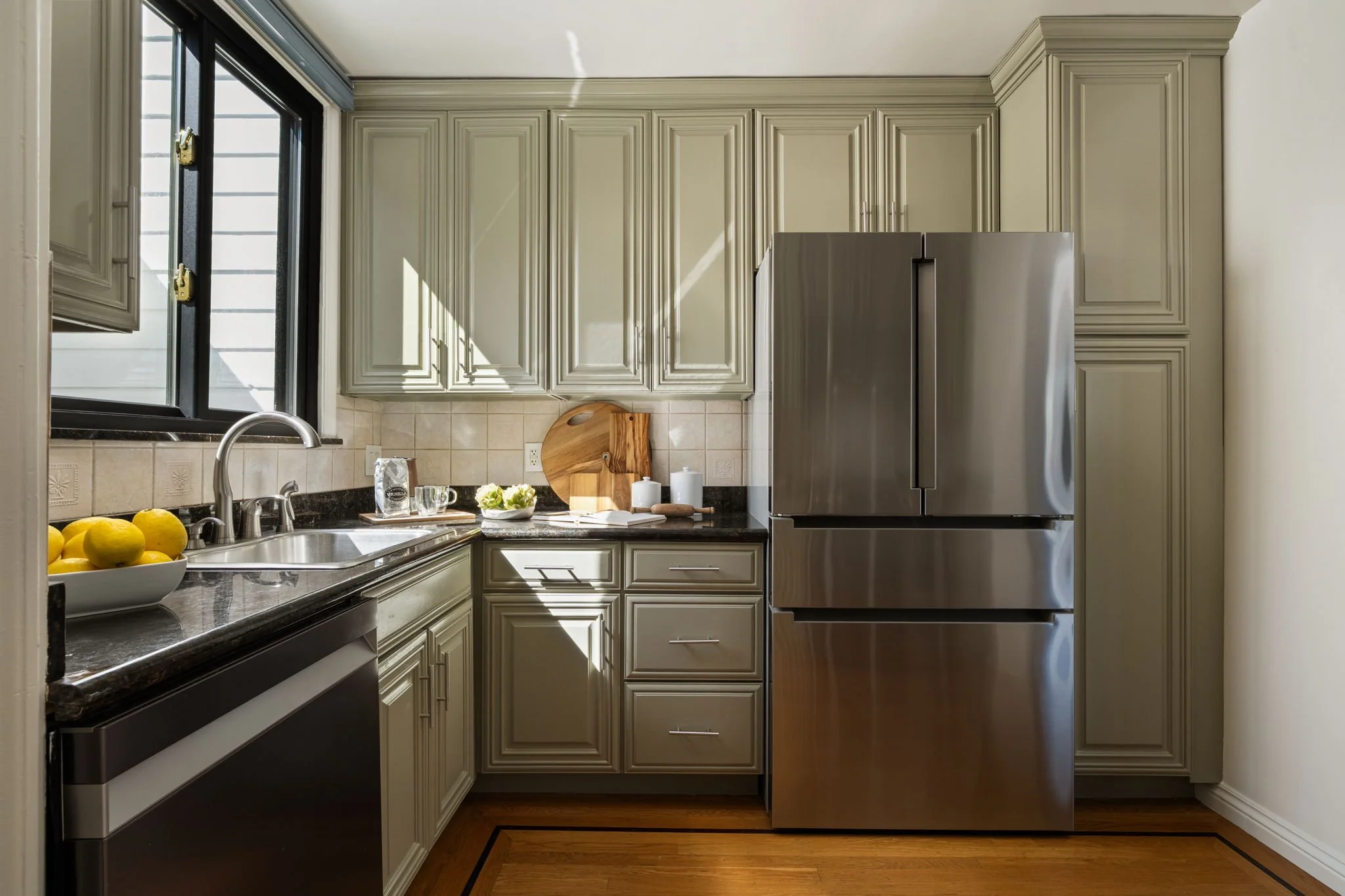 Kitchen with beige cabinets, stainless steel refrigerator, black granite countertop, and a window with sunlight. A bowl of lemons on the counter, and dishes near the sink.