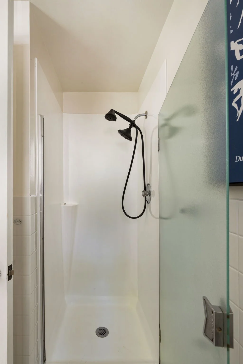 Empty shower with a black handheld showerhead on a white shower wall, glass door on the right, and white tiled walls on the left.