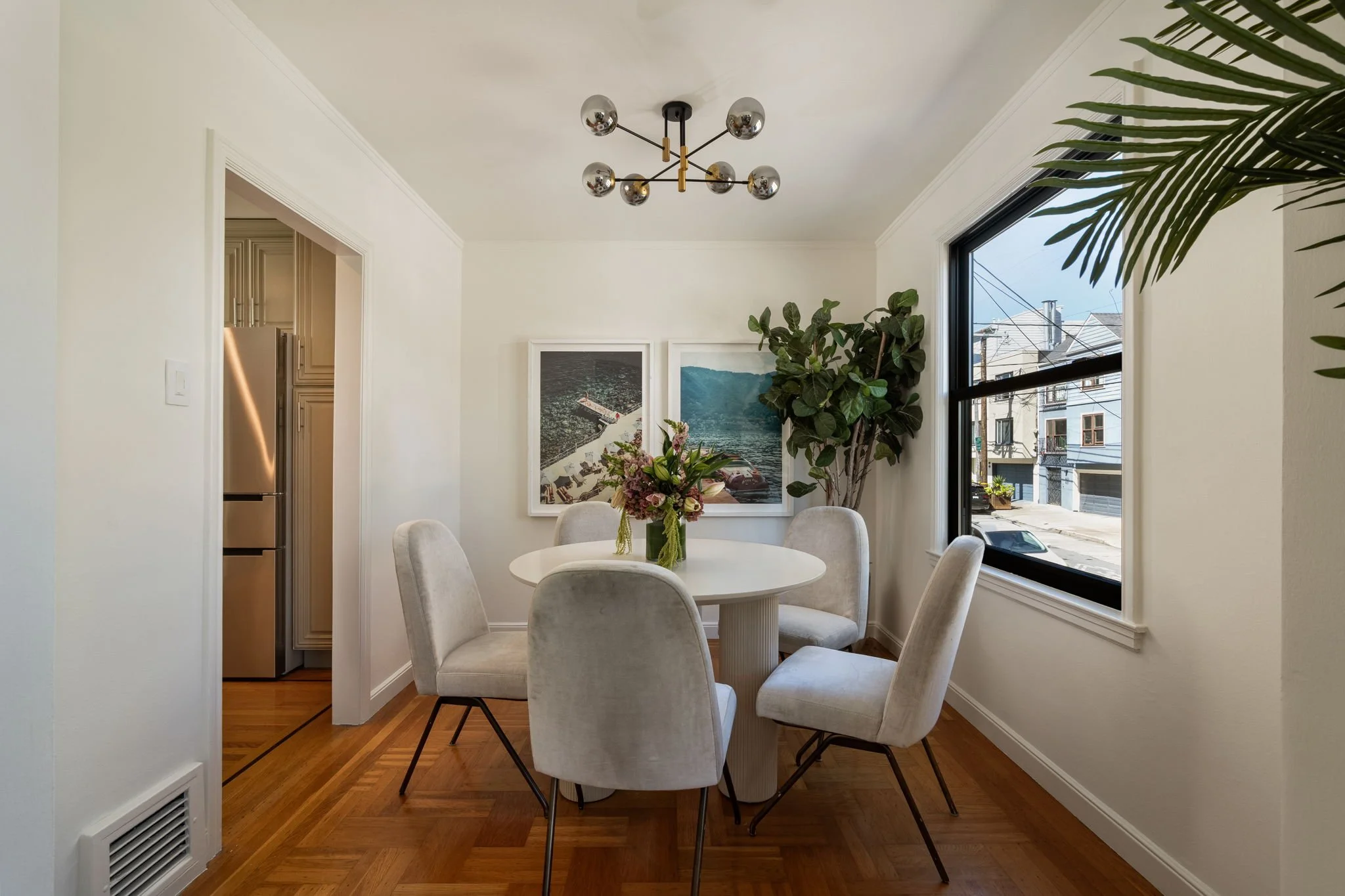 Dining room with a white round table and beige upholstered chairs, a floral centerpiece, large window with city view, and modern chandelier overhead.