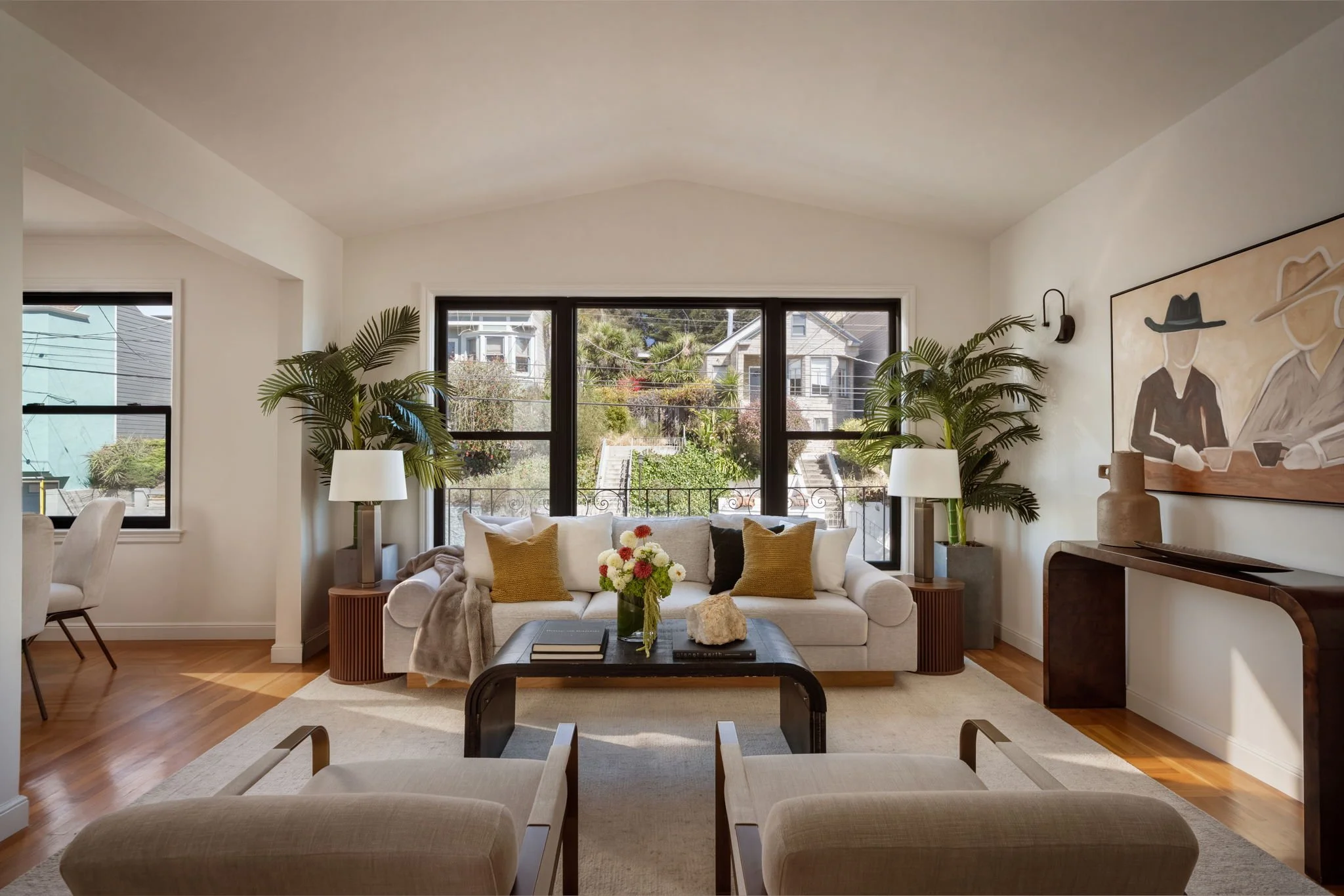 A bright living room with large windows overlooking a neighborhood street, with glass doors at the back, a white sofa with yellow pillows, a black coffee table with a vase of flowers, two beige armchairs, and indoor plants flanking the sofa, with a large abstract painting on the wall.