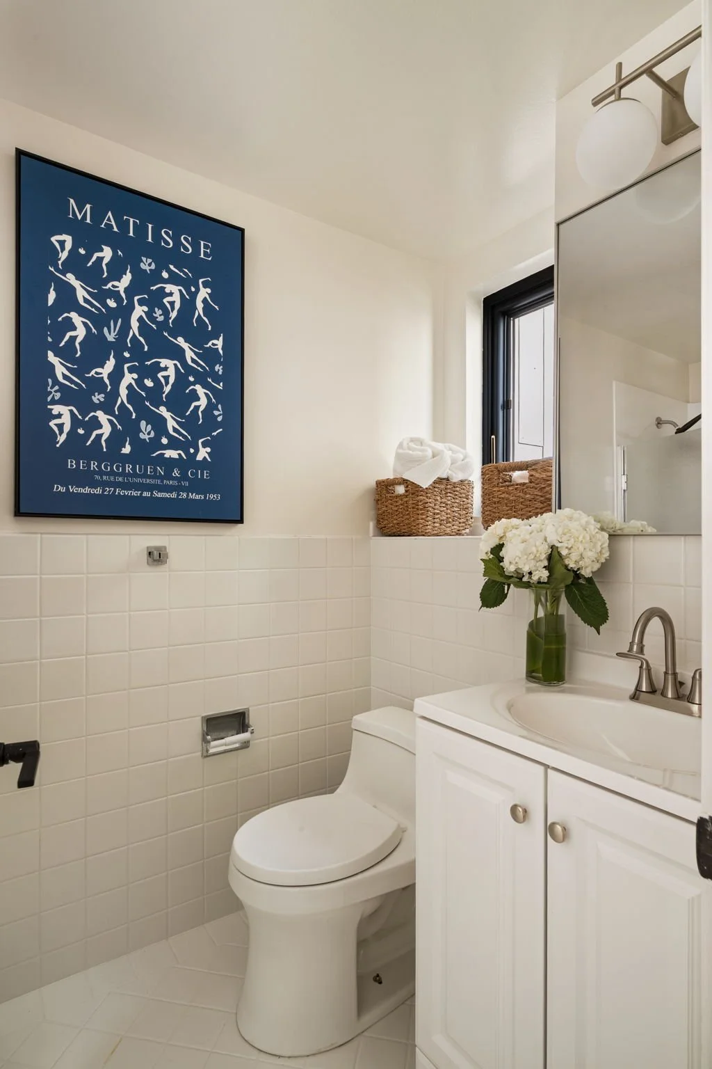 Small bathroom with white tile walls, a white toilet, a white vanity with a sink, a vase with white hydrangeas on the counter, a framed Matisse art print on the wall, a window with a black frame, and baskets on a ledge.