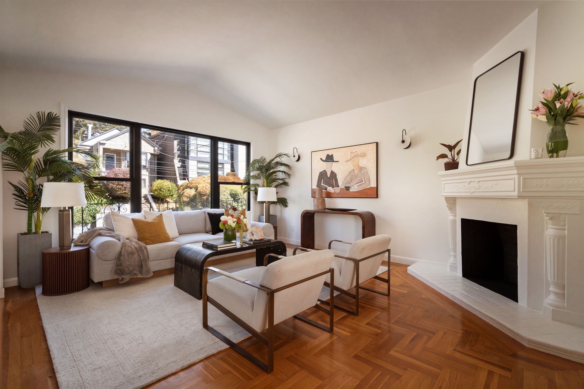 A living room with a white sofa, beige armchairs, and a black coffee table with flowers on it. Large windows show a view of neighboring houses and greenery. There is a fireplace with a mirror, a painting of two women, and various plants and lamps for decoration.