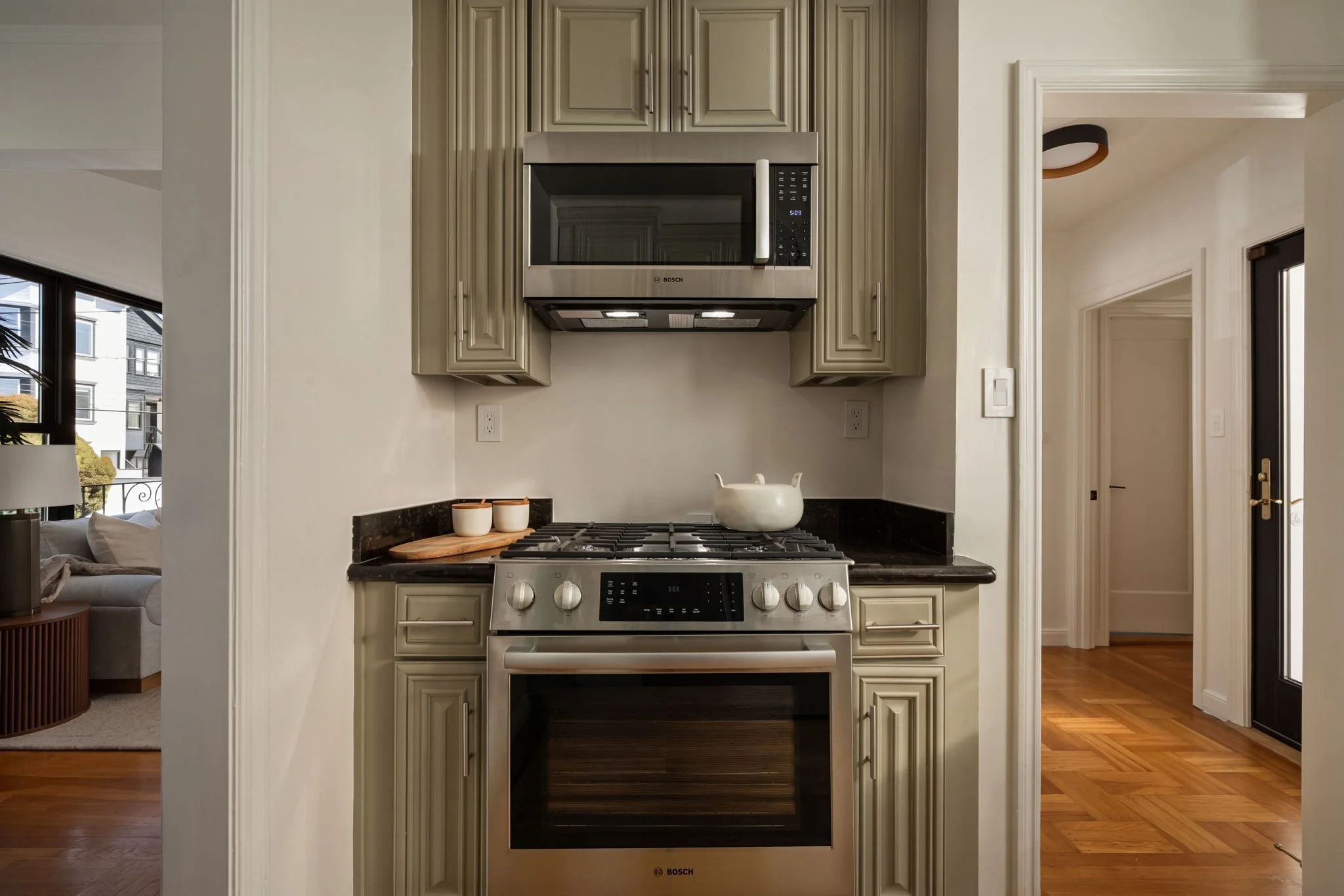 Stainless steel Bosch microwave mounted above a stove in a kitchen with beige cabinets and black countertops.