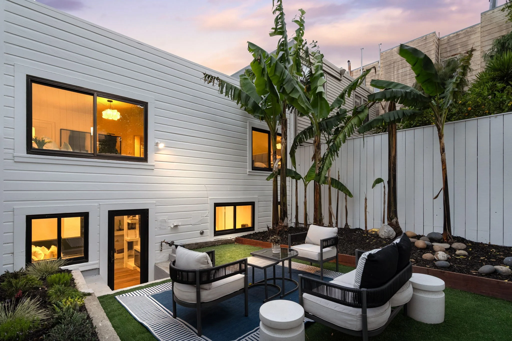 Modern backyard patio with white house exterior, large green banana trees, outdoor seating with cushioned armchairs, black patio tables, striped outdoor rug, and a small patch of artificial grass at sunset.