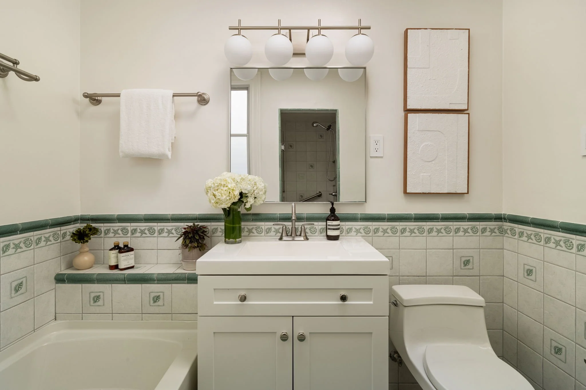 A bathroom with a large mirror above a white vanity, a vase of white flowers, soap, and bathroom products, with decorative tiles and framed art on the wall, a towel rack with a white towel, and a view into a shower area.