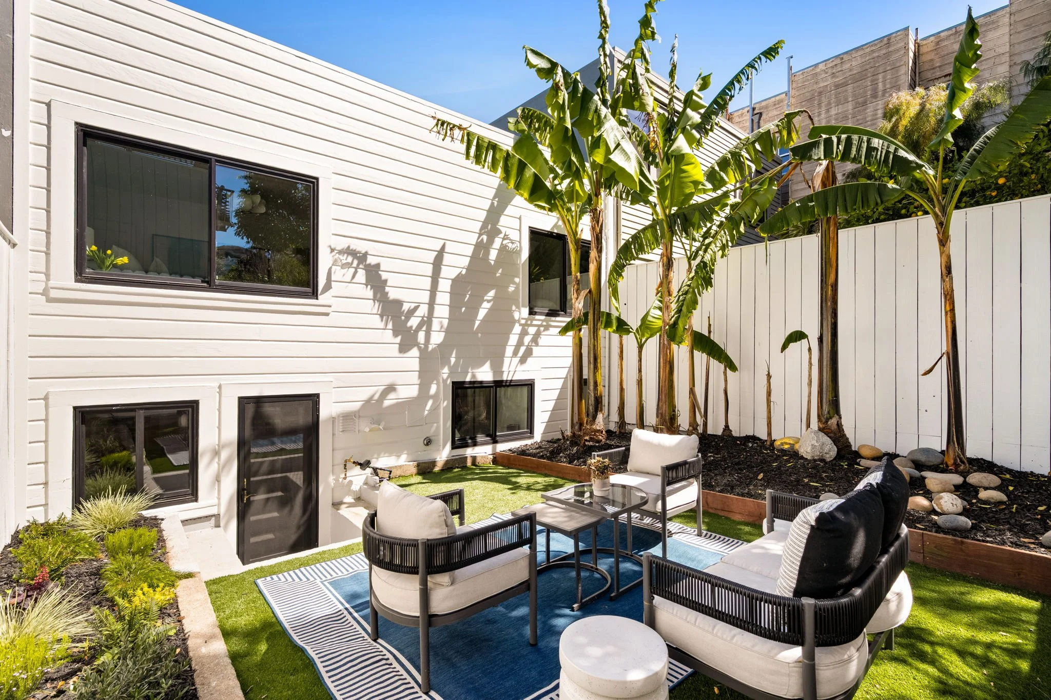 A backyard patio with black and white outdoor furniture, a blue and white striped rug, and several banana trees against a white house with black framed windows and a white fence.