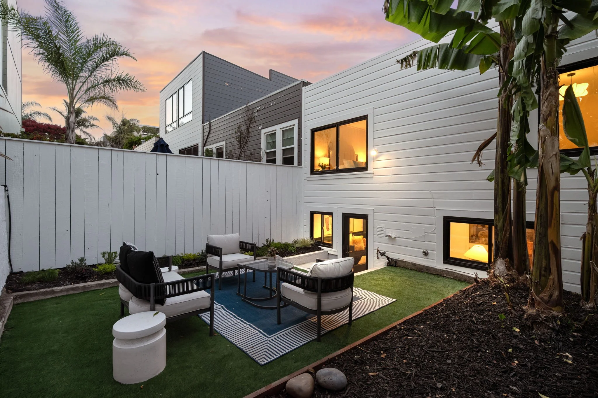A backyard patio with outdoor furniture including chairs and a table, surrounded by a garden with palm trees and a white fence, with a modern white and gray house in the background during sunset.