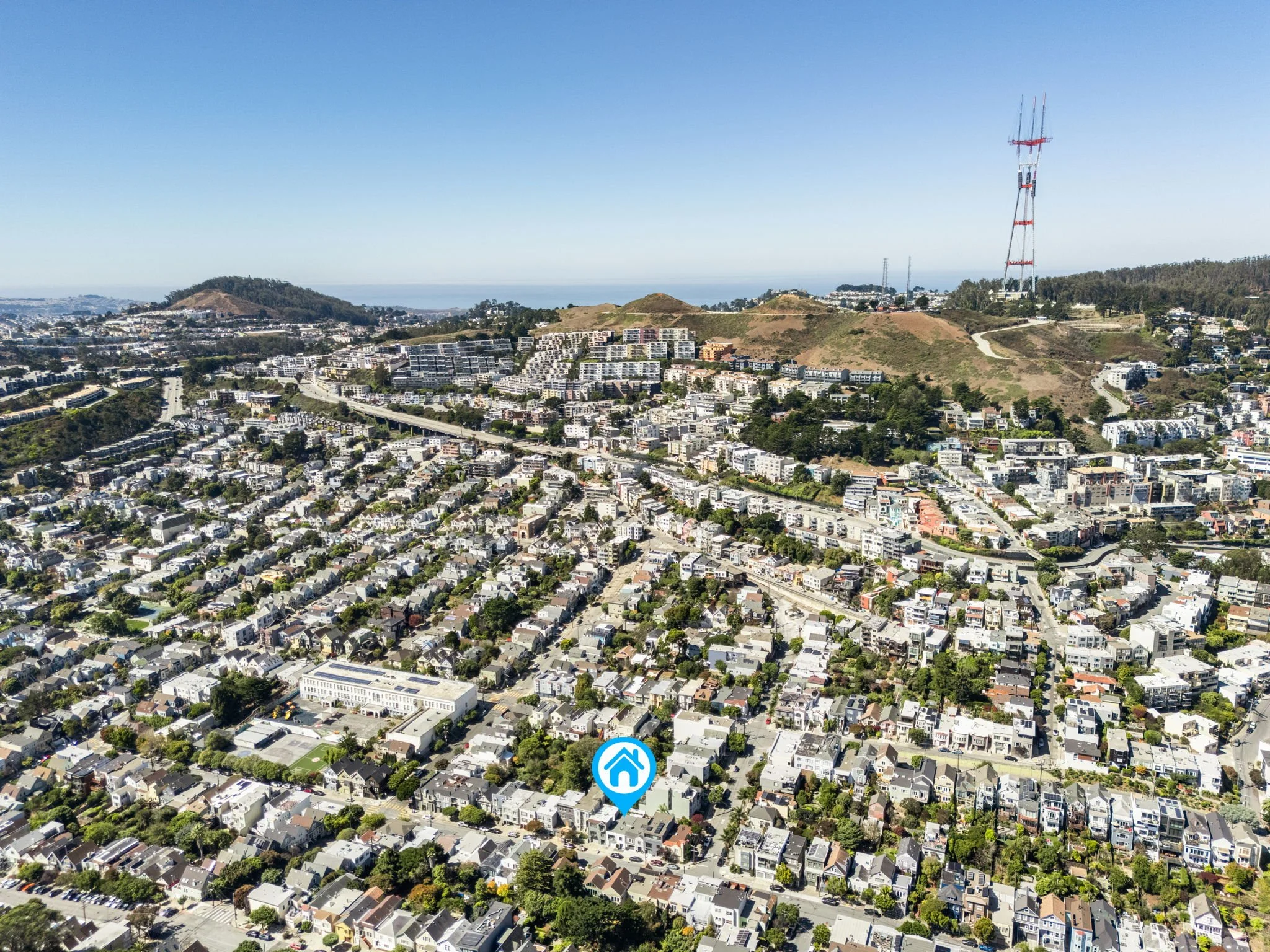 Aerial view of a cityscape with a hillside, numerous residential buildings, and a large red and white communication tower in the distance.
