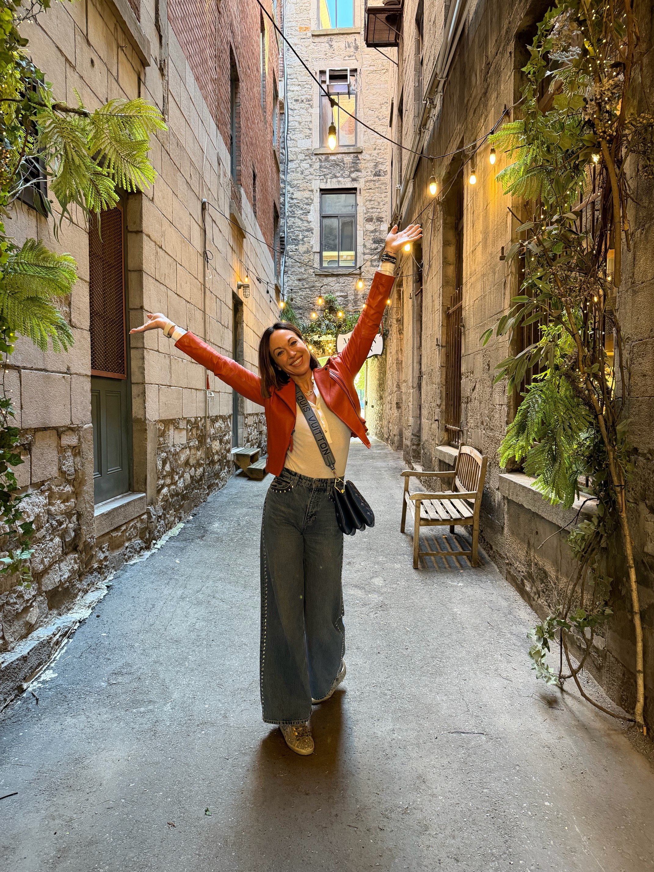 Smiling woman in a red leather jacket and high-waisted jeans standing with arms raised in an alleyway decorated with string lights and greenery, surrounded by stone and brick buildings.