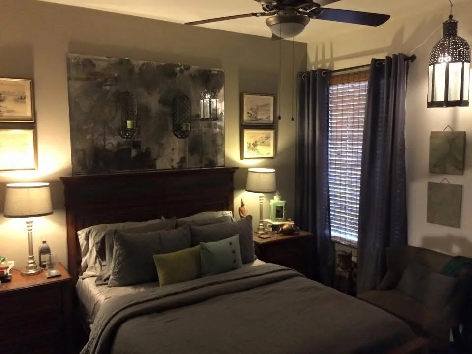 A cozy bedroom with a bed, pillows, nightstands with lamps, a ceiling fan, windows with blinds and curtains, a small sofa, and wall art.