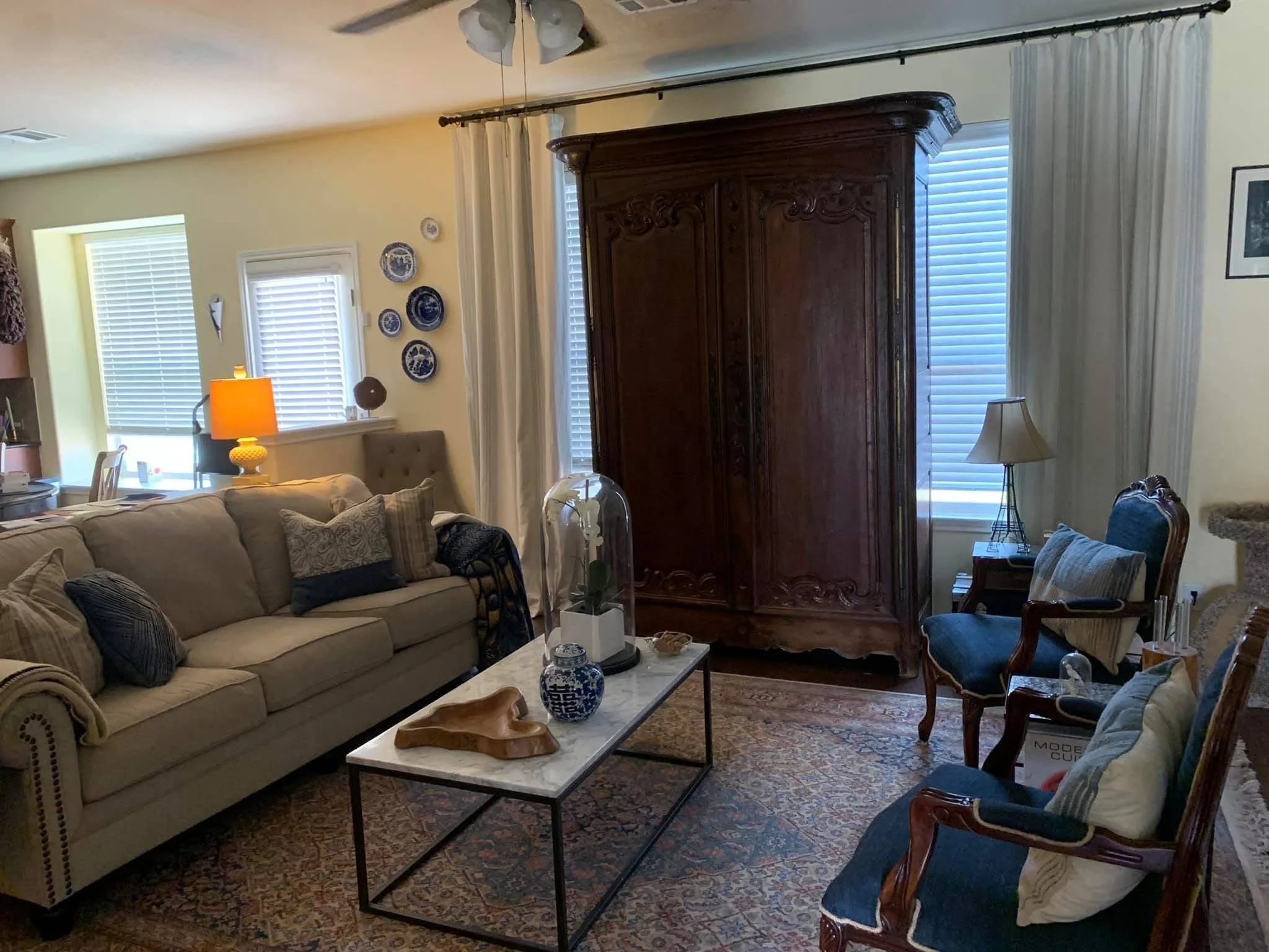 A cozy living room with a beige sofa with patterned pillows, a marble-top coffee table with decorative vases and a wooden sculpture, a vintage wooden armoire, a small table with a lamp, and several windows with white blinds and curtains.