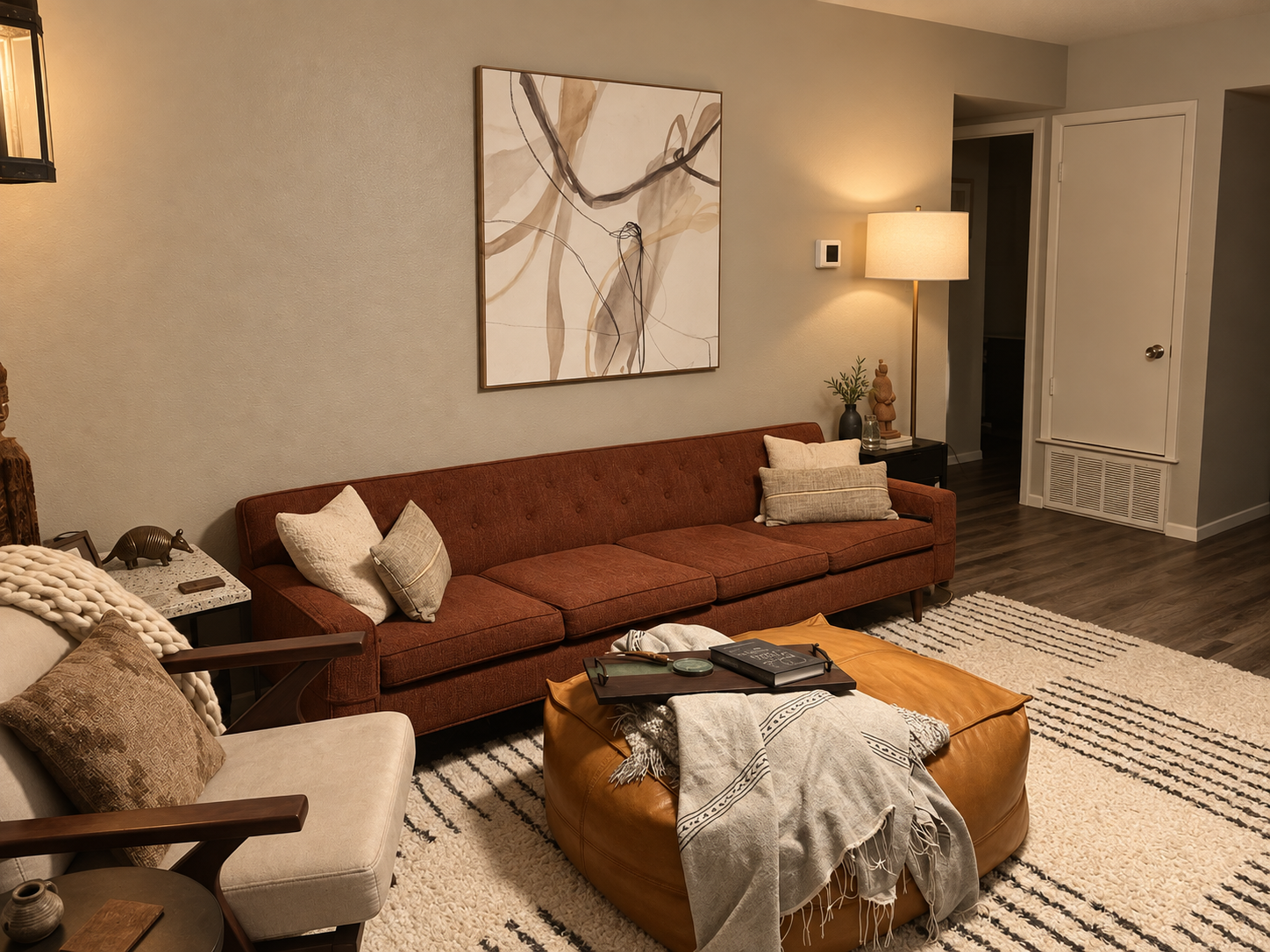 Living room with a rust-colored sofa, a beige armchair, a coffee table, a floor lamp, wall art, and decorative pillows and blankets.