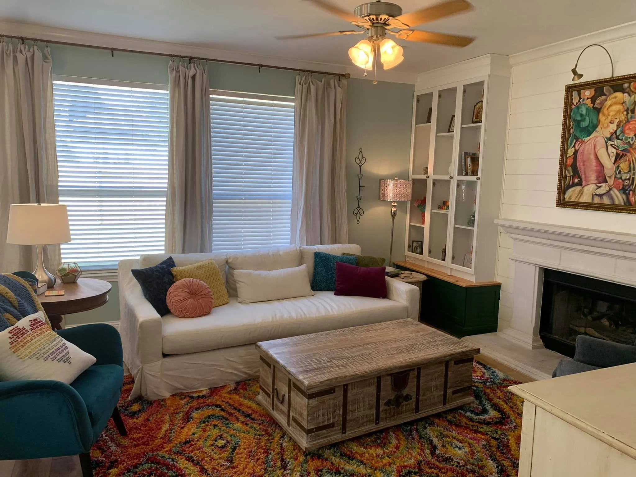 Living room with large white sofa with colorful pillows, a blue armchair with a cross-stitched pillow, a wooden coffee table, a fireplace, a white custom built-ins, and bespoke reproduction painting.