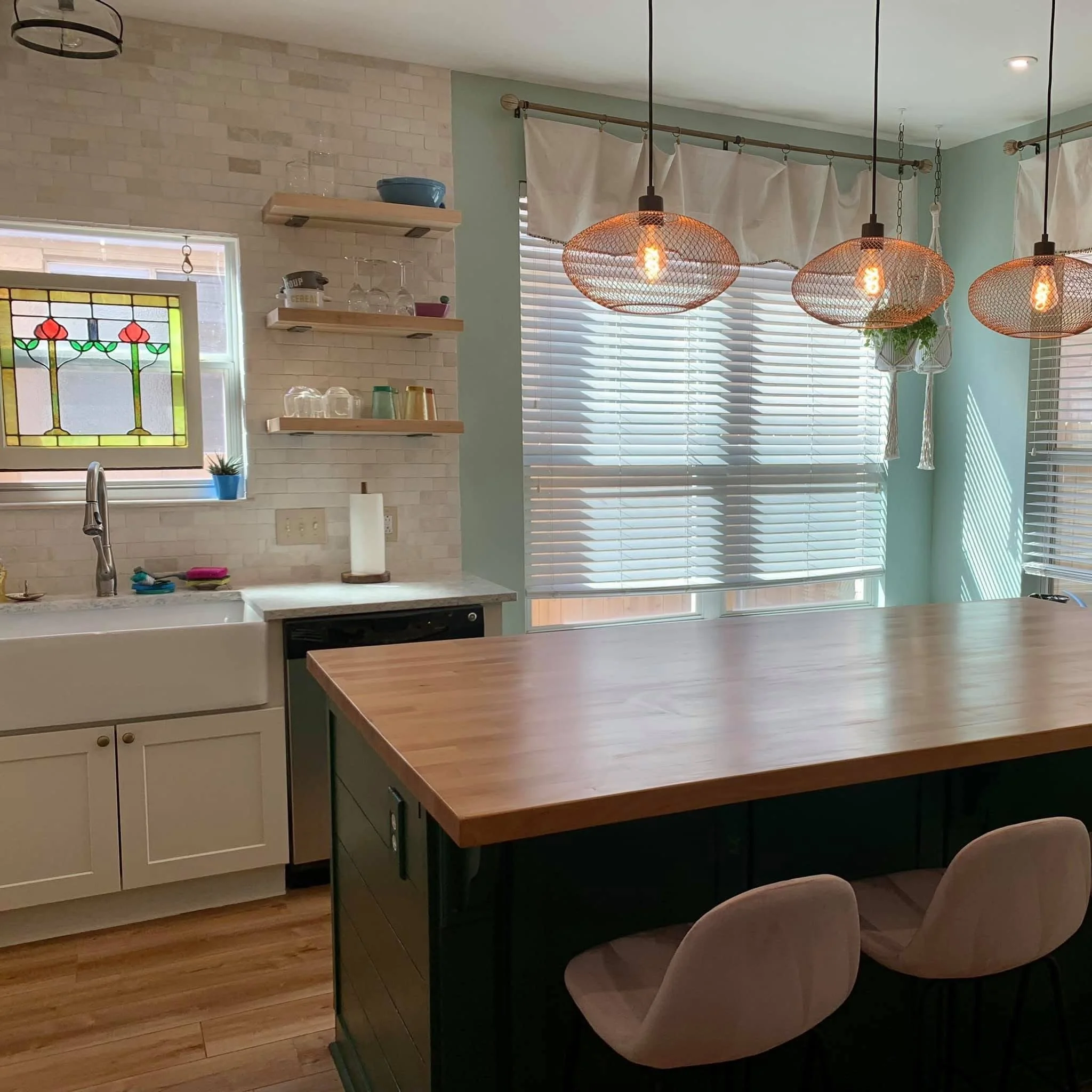 Modern kitchen with limestone tile backsplash, off-white cabinets, and wooden open shelves holding glassware and bowls. A window with stained glass design, and three pendant lights with copper mesh shades hanging above a butcher block island.