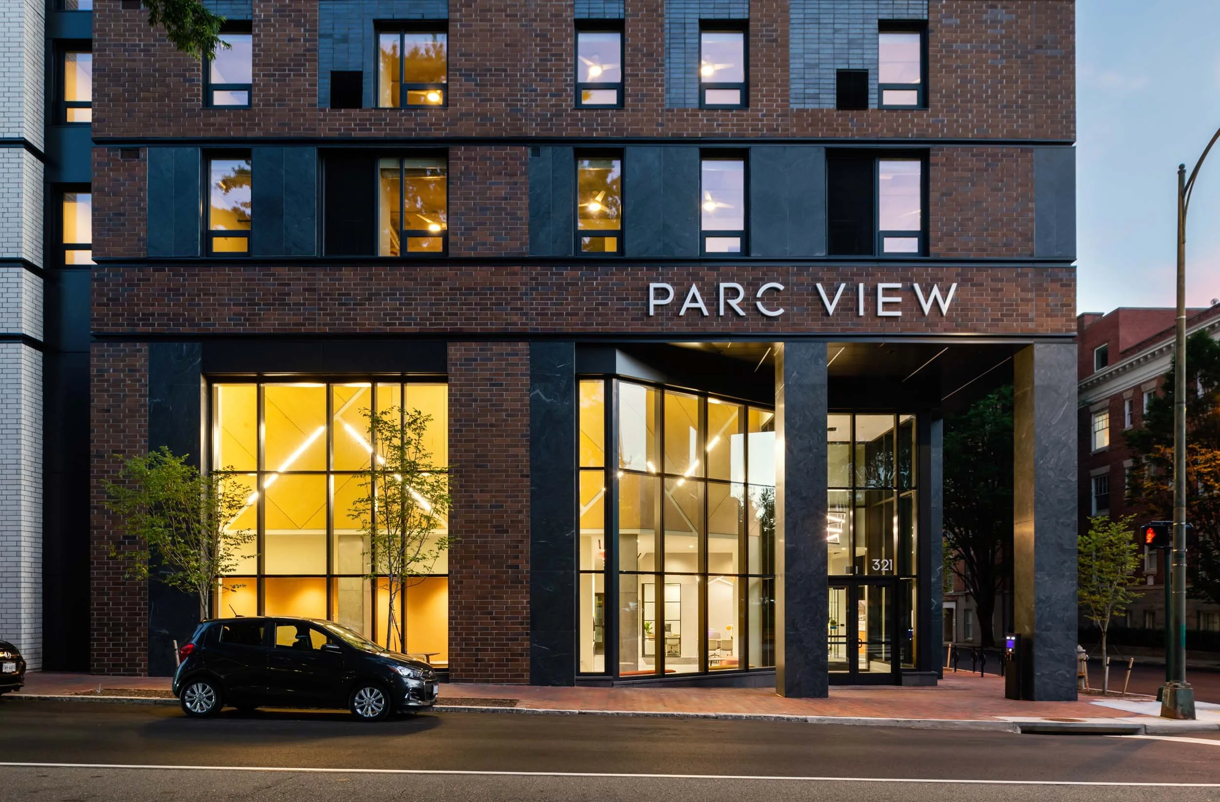 A modern multi-story building with a brick facade, large glass windows, and illuminated interior lights. The sign on the building reads "PARC VIEW." There is a black car parked in front and some small trees on the sidewalk.
